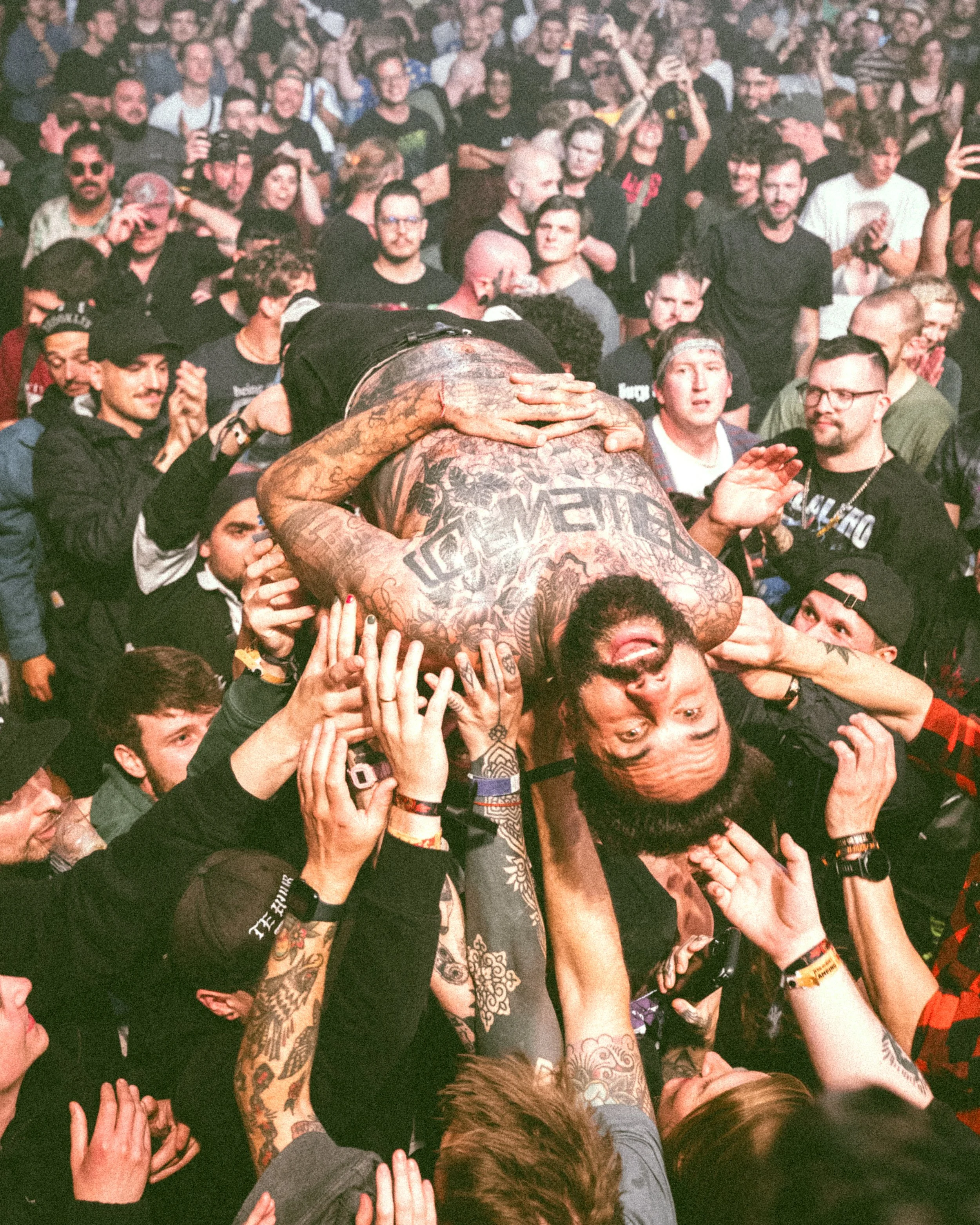 Crowd at a concert or festival holding up a large tattooed man in the air, who appears to be crowd surfing, with the audience supporting him with their hands.
