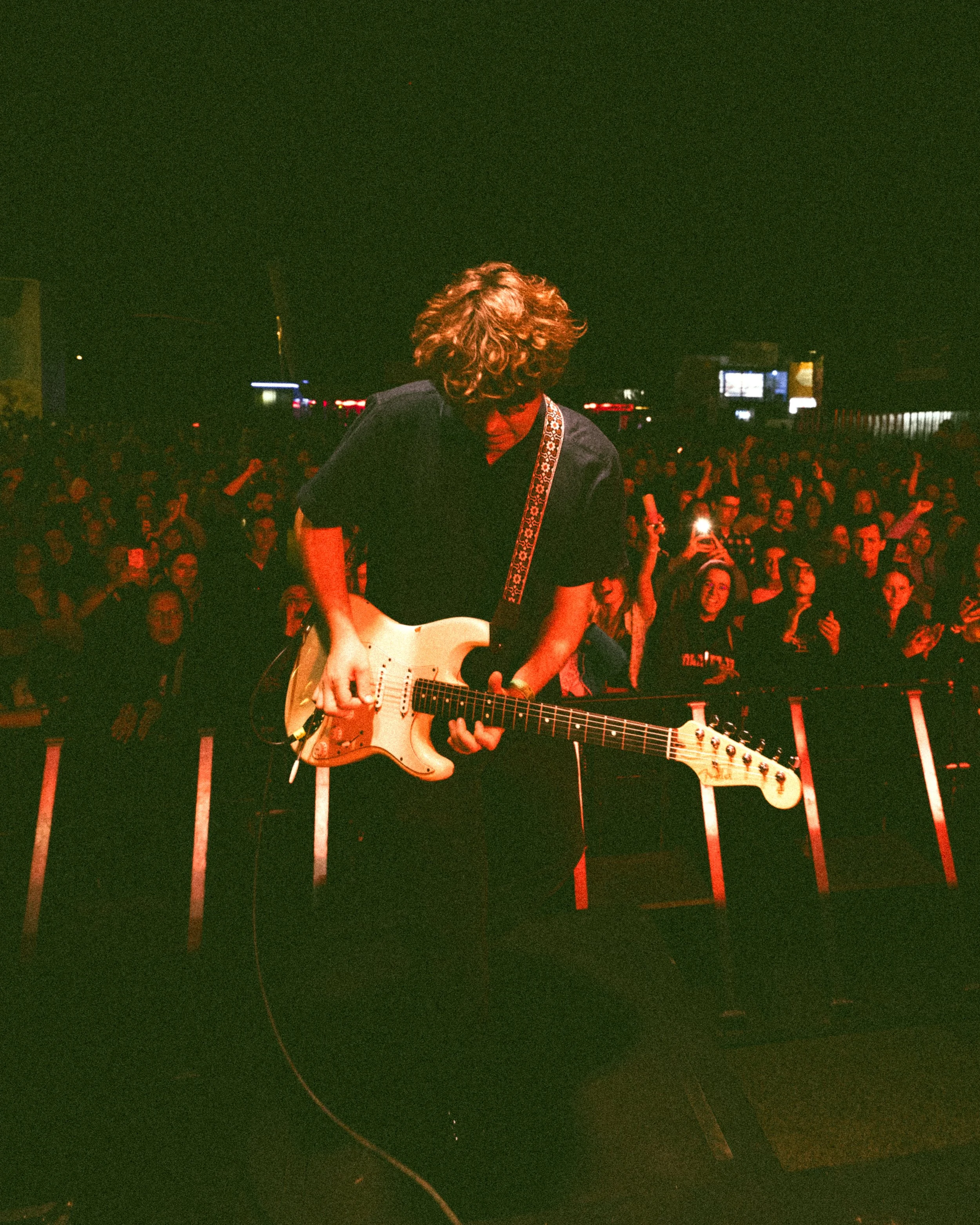 A young musician with curly hair playing an electric guitar on stage at a concert, with an audiences in the background.