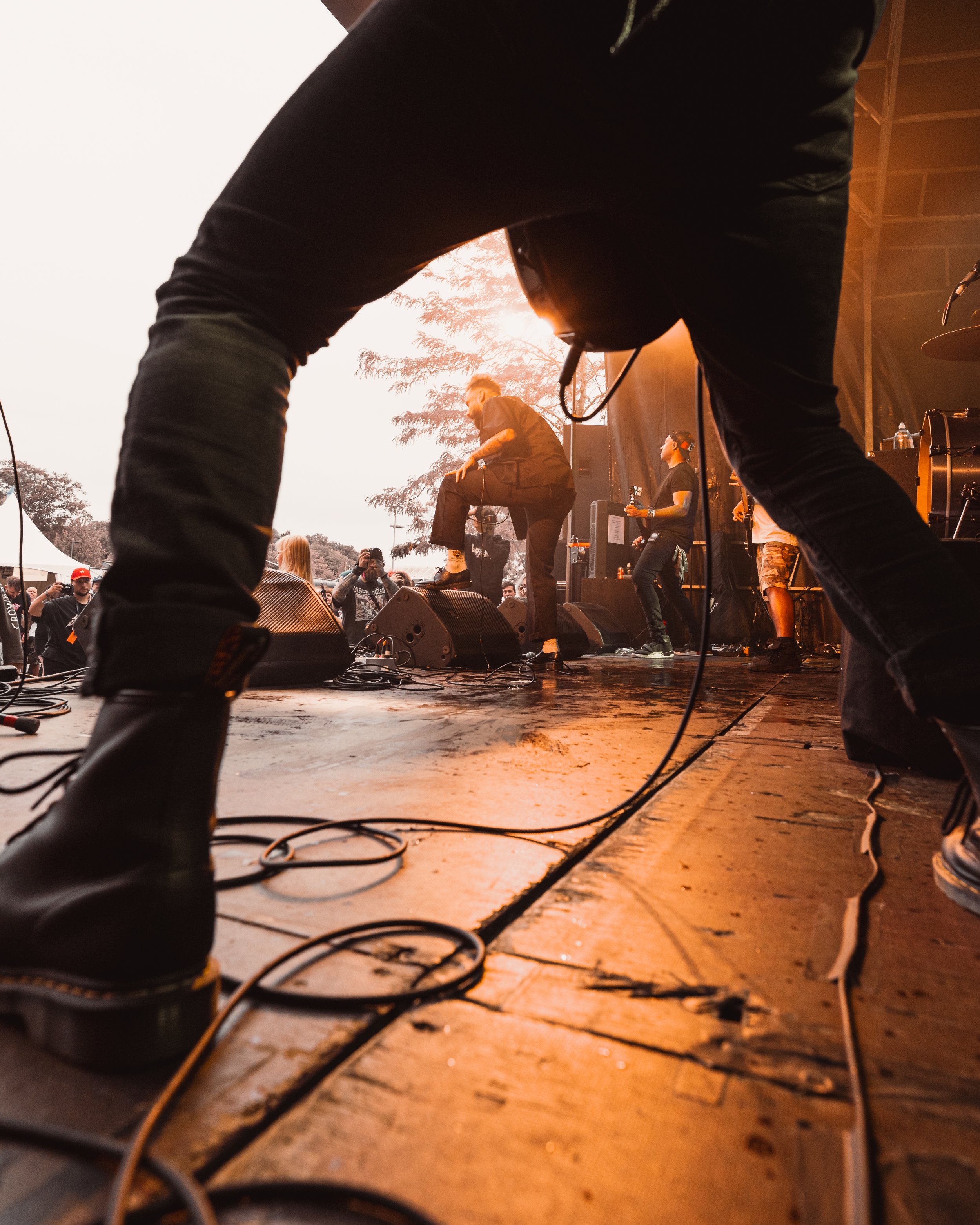 View from the stage at a concert, showing performers and an audience outside. The photo is taken from a low angle, focusing on the edge of the stage and equipment with concertgoers in the background.