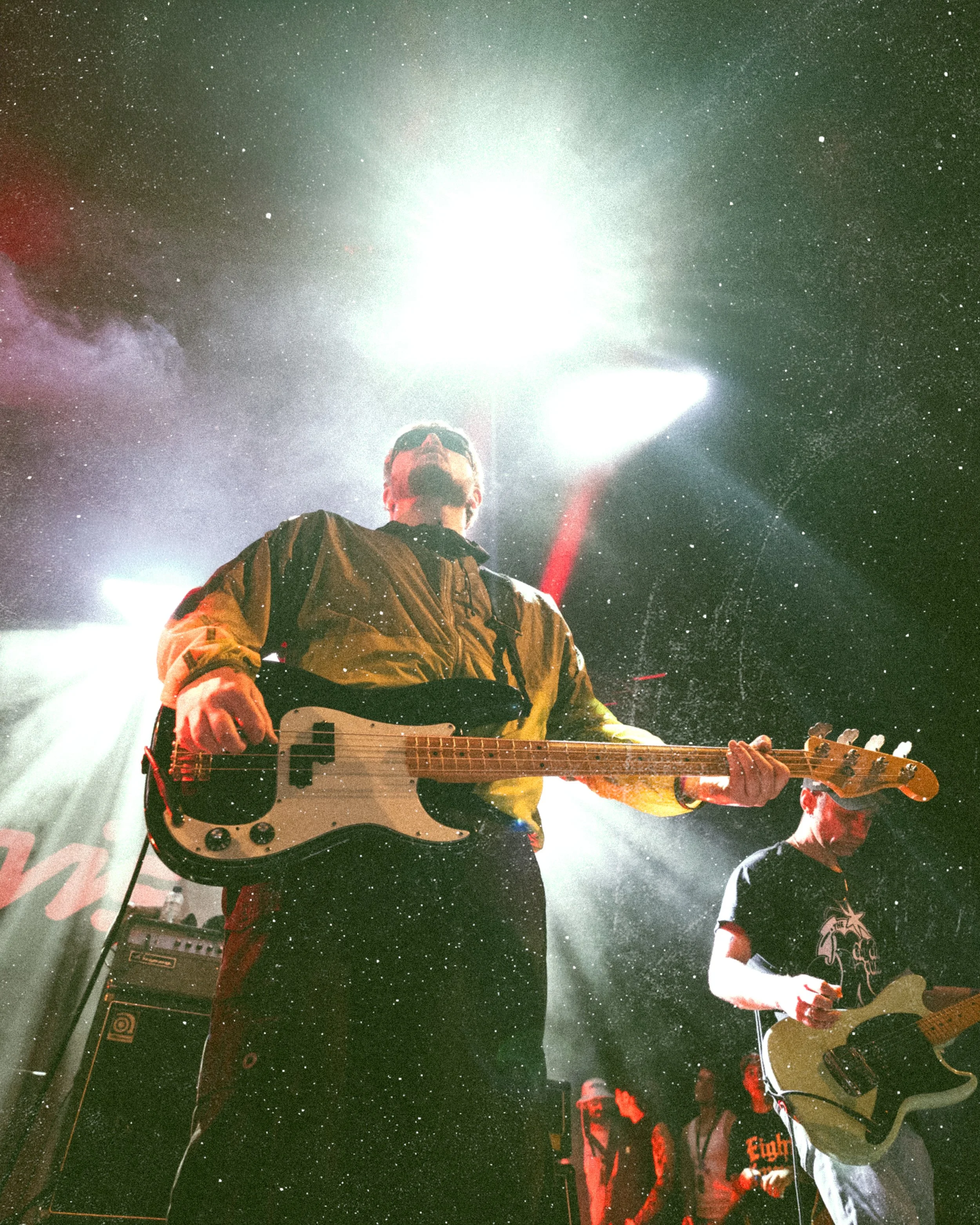 A musician playing guitar on stage with bright stage lights and smoke effects, a second guitarist visible in the background.