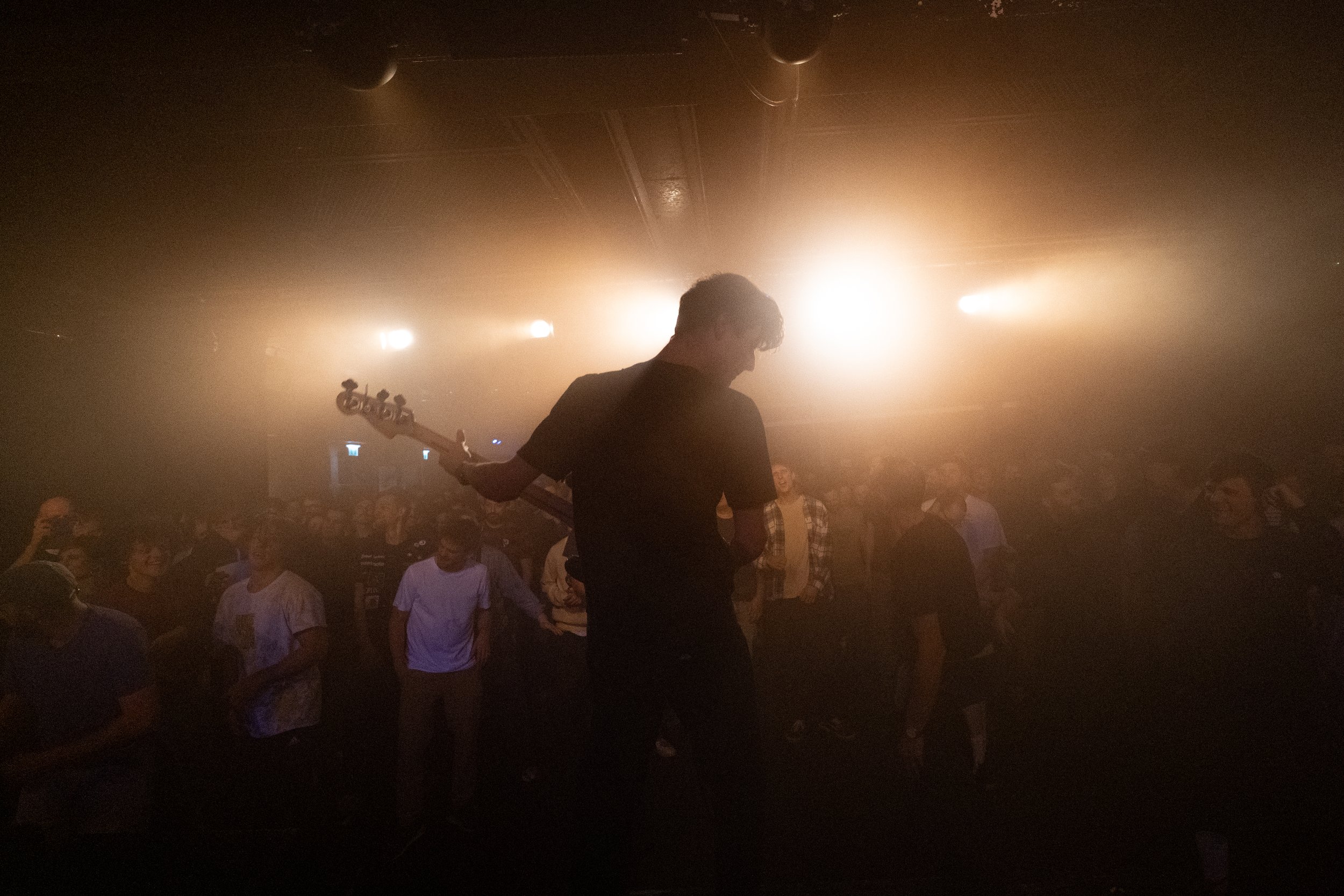 A musician playing an electric guitar on stage, backlit by bright stage lights, with an audience watching in a dimly lit concert venue.