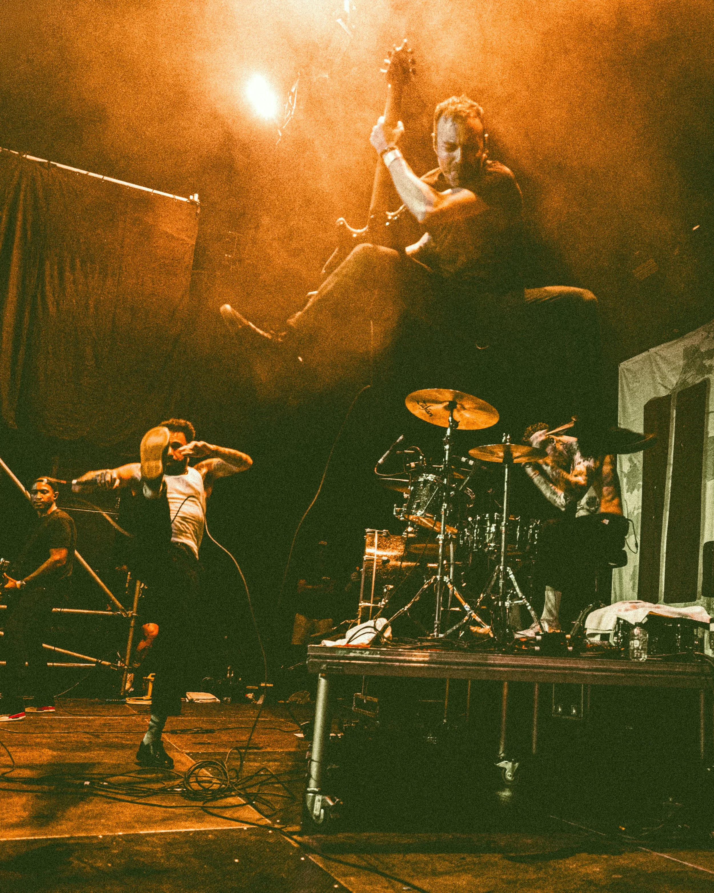 Musicians performing on a stage with dramatic lighting, with one musician jumping in the air and others playing instruments.