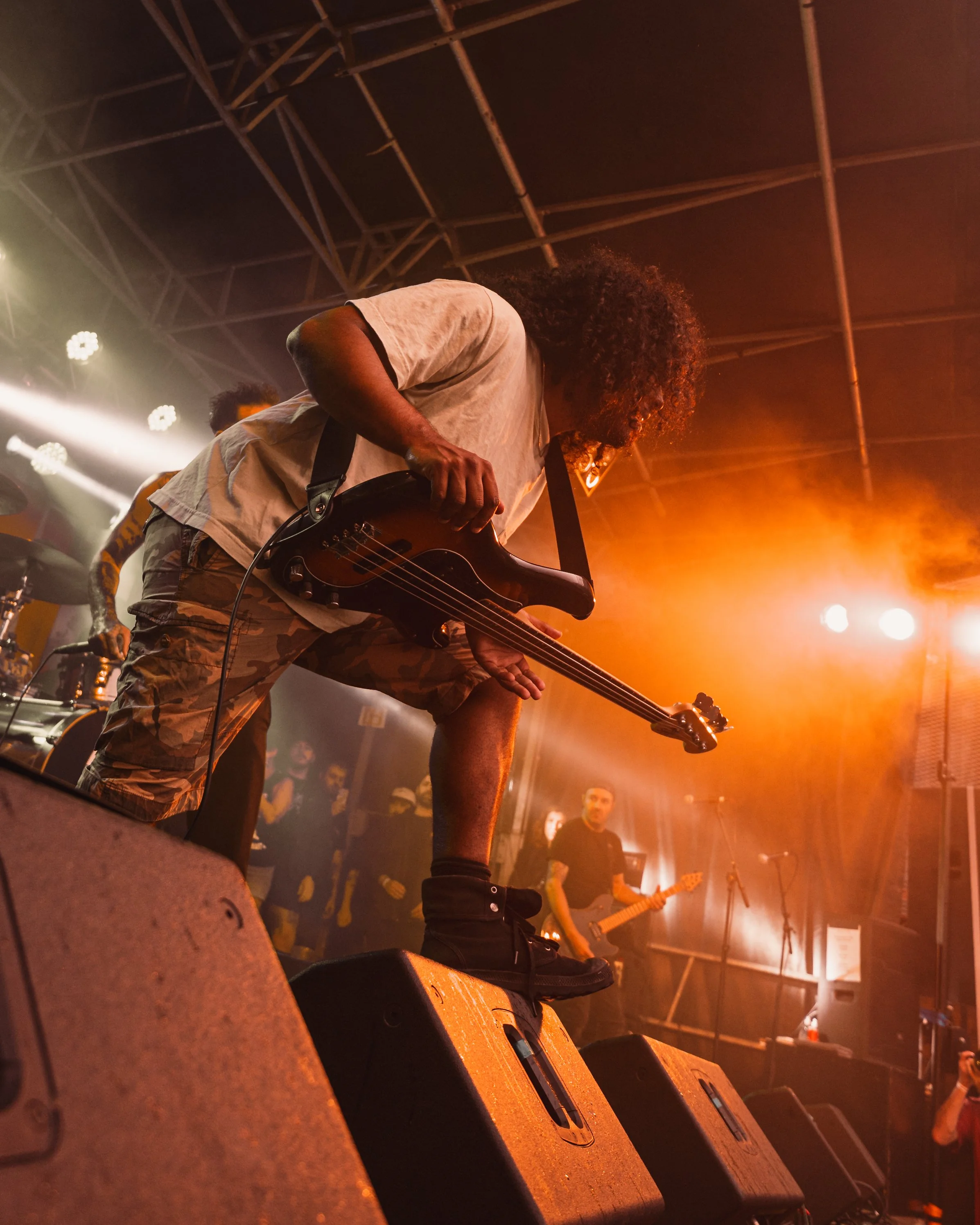 A musician playing an electric bass guitar on stage with orange lighting and an audience in the background.