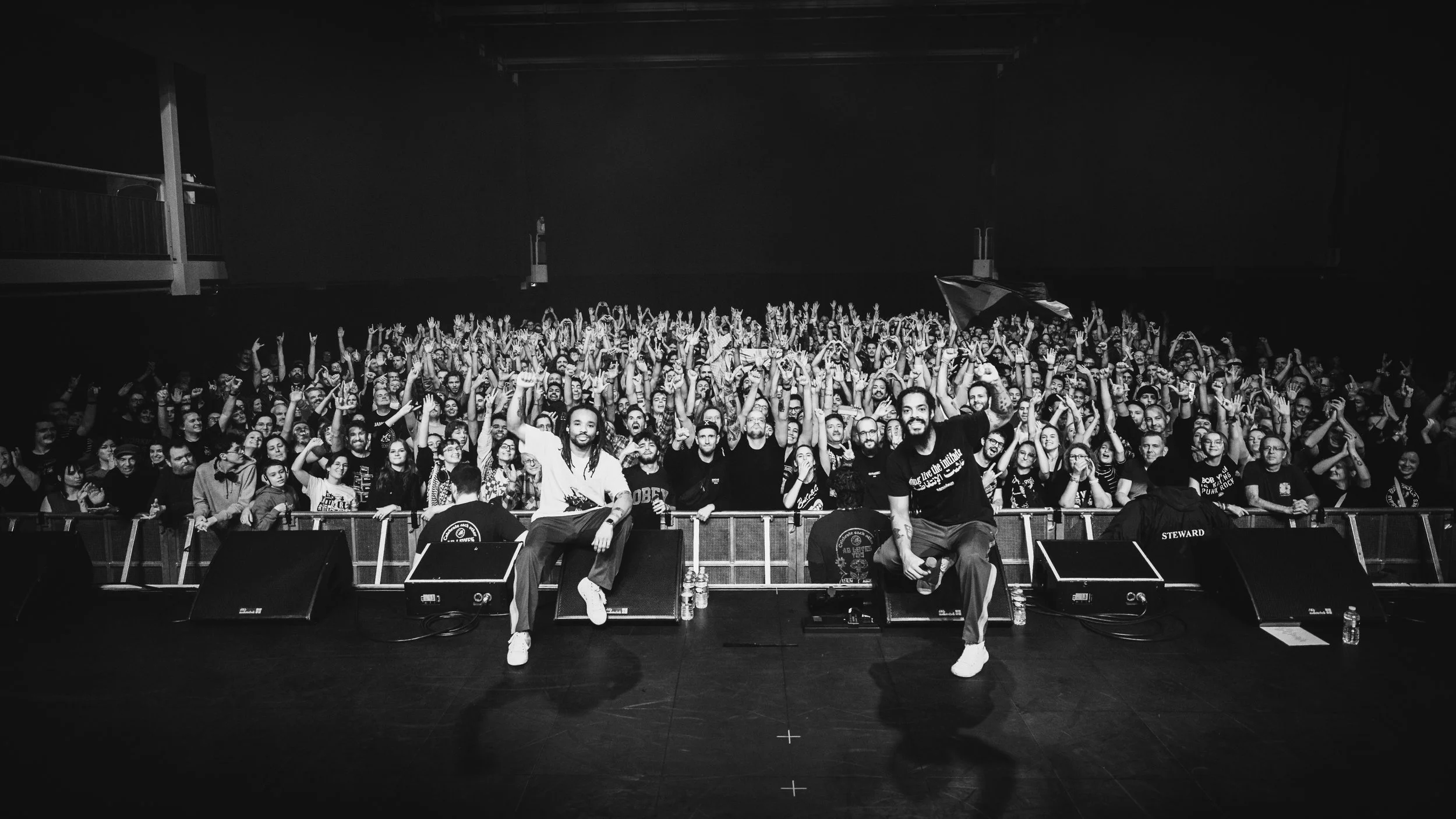 Black and white photograph of a large crowd at a concert, with three performers sitting on the stage in the foreground, facing the camera and waving. The audience is enthusiastic with many raising their hands and cheering.
