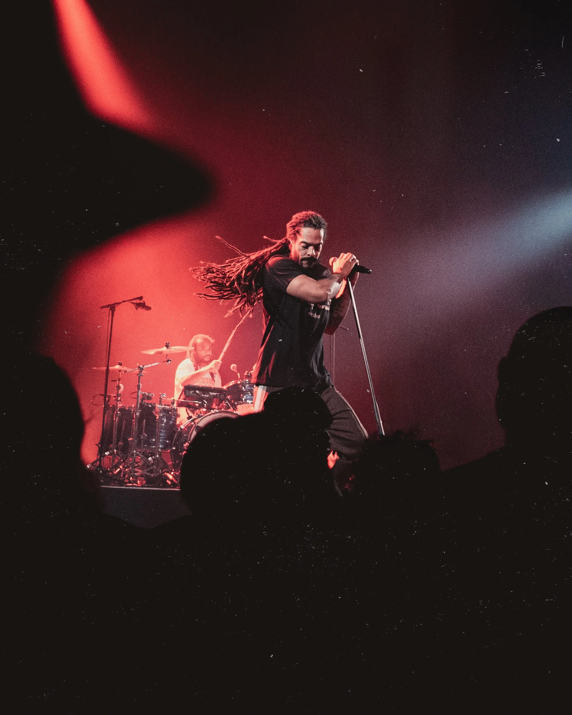 A male singer with dreadlocks performing on stage, with a drummer playing in the background, under red stage lighting.
