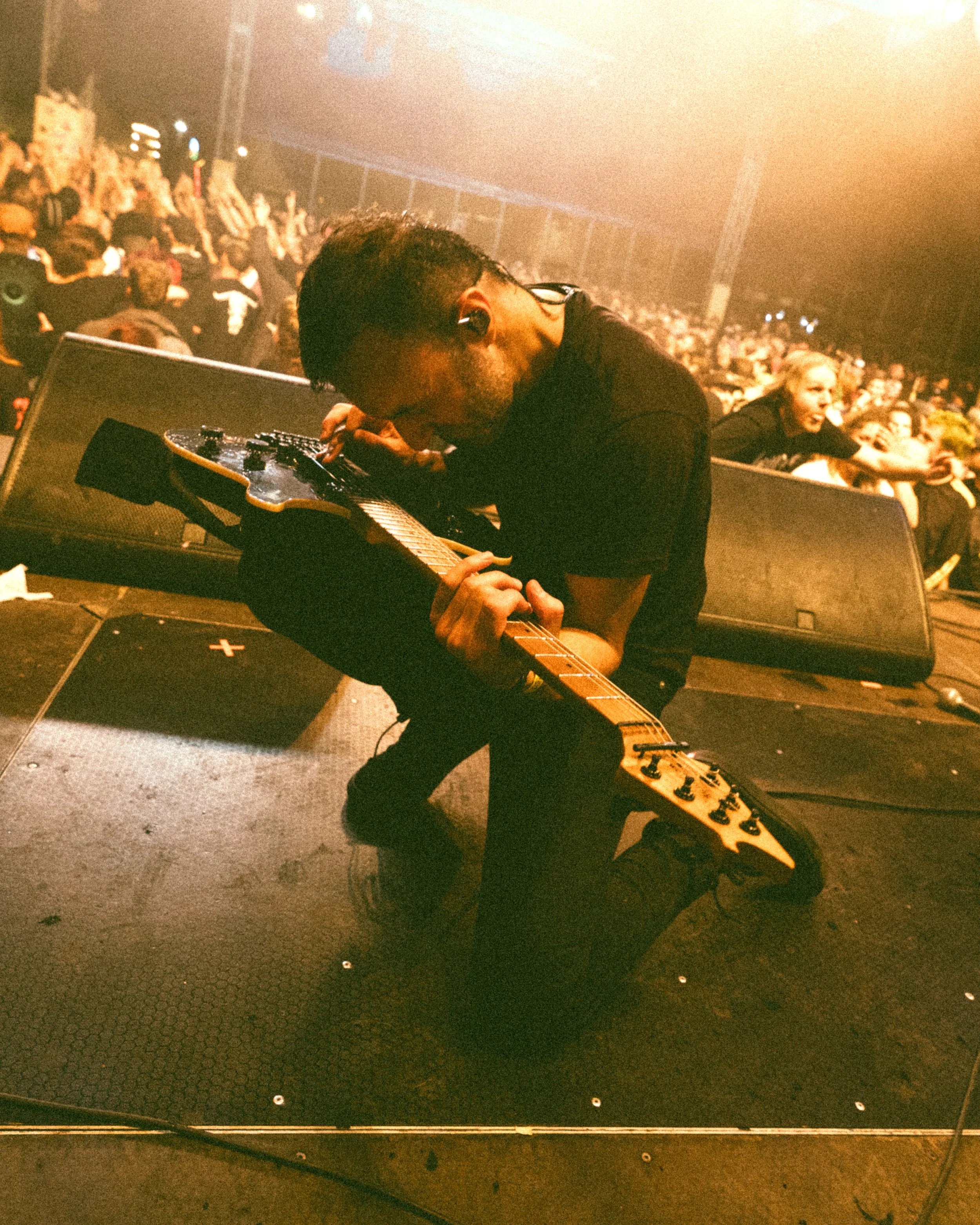 Kneeling male guitarist adjusting guitar at a live concert with crowd in the background.