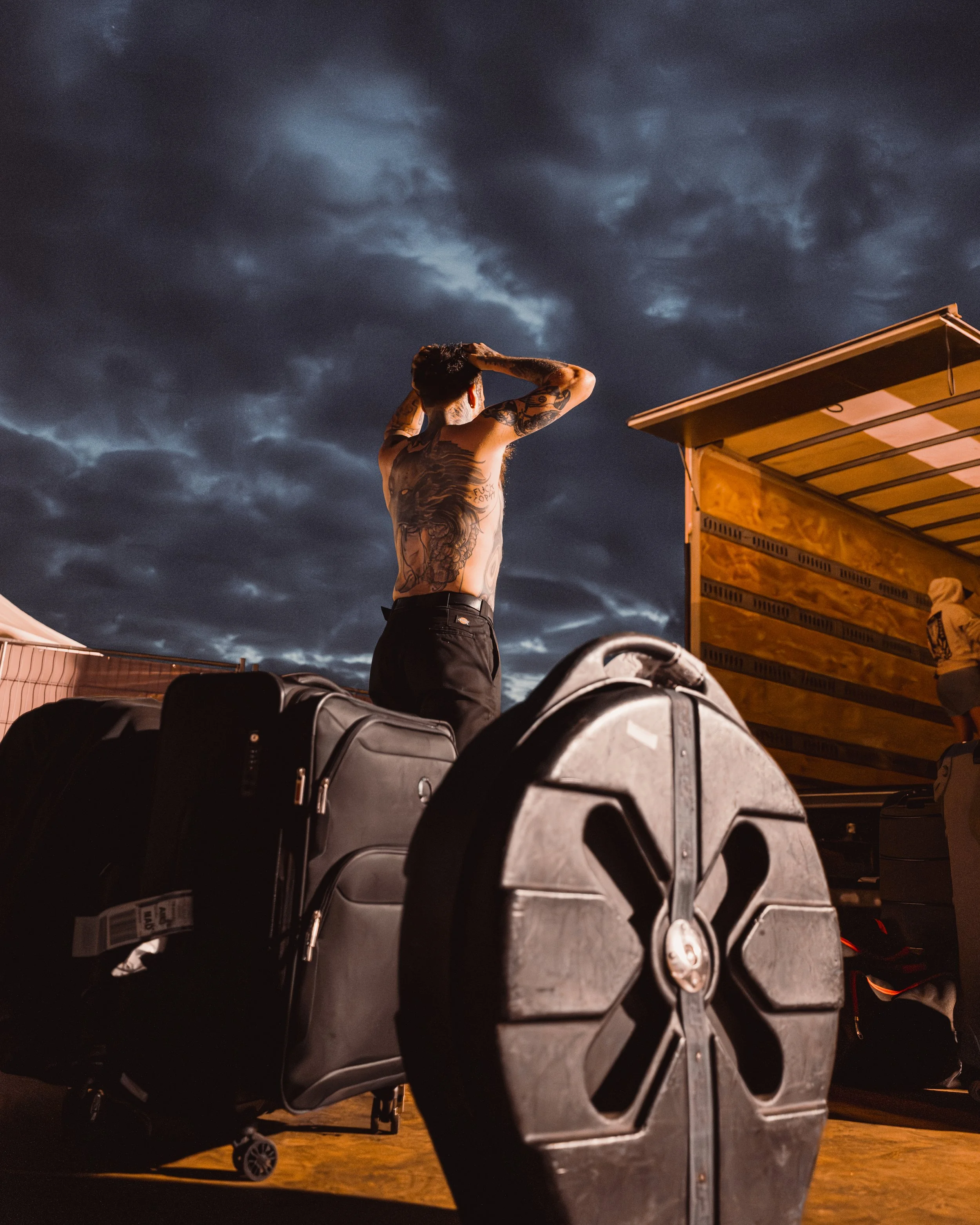 A shirtless man with tattoos stands outdoors at night, adjusting his hair against a dark, cloudy sky, with luggage and a truck nearby.