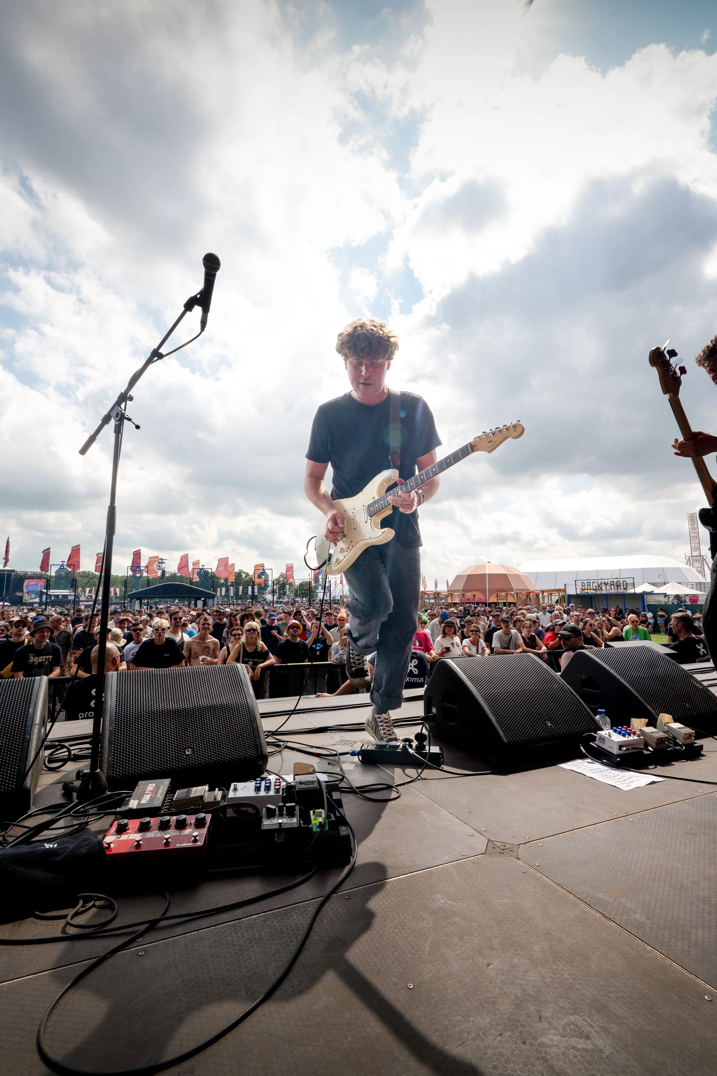 A musician playing an electric guitar on an outdoor stage at a concert with a crowd in front and cloudy sky above.