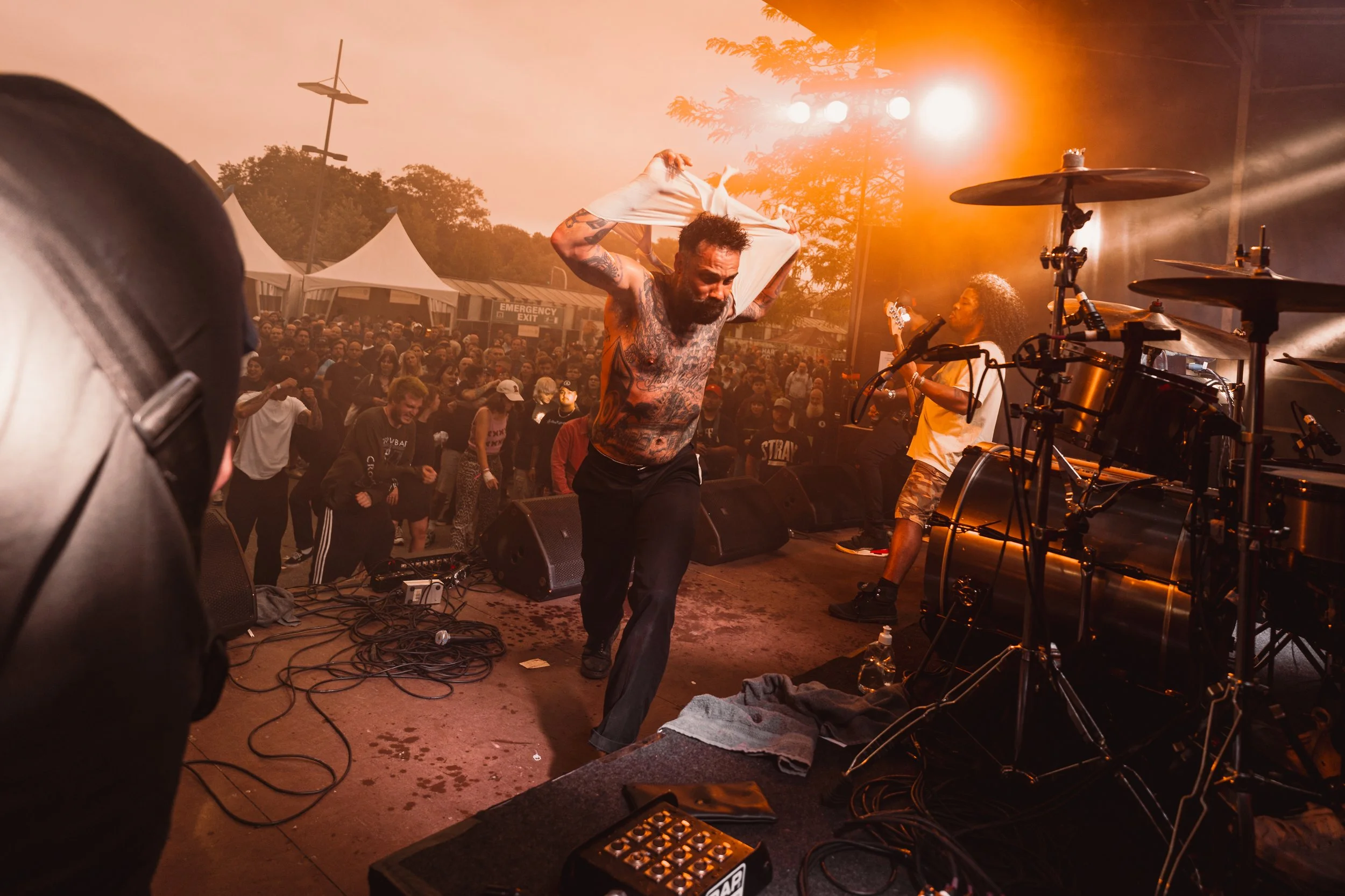 Musicians performing on stage at an outdoor concert, with a shirtless man with tattoos dancing and a woman singing beside him, stage equipment and an audience in the background.
