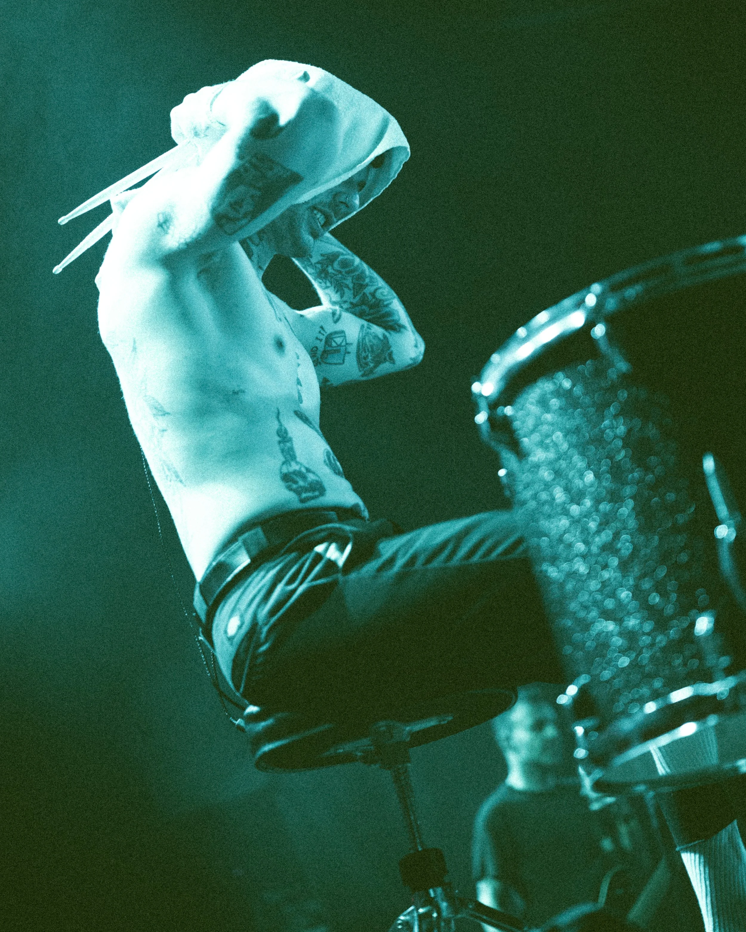 A shirtless man with tattoos on his arms and torso, wearing a hoodie over his head, sitting on a drum stool with a drum set in front of him, in a dimly lit room with green lighting.
