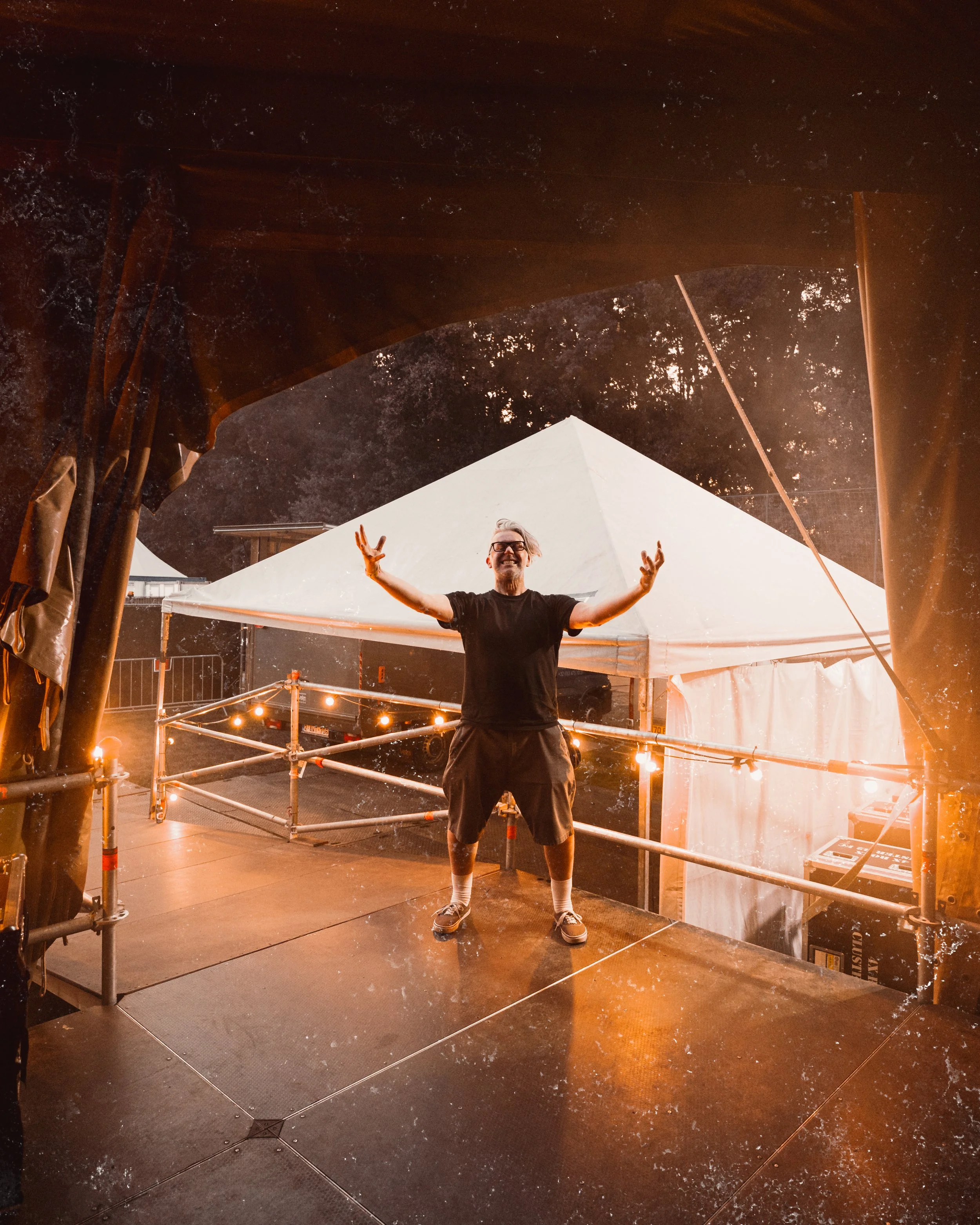 A man standing on a stage with arms outstretched, smiling, during an outdoor event at night, with tents and string lights in the background.