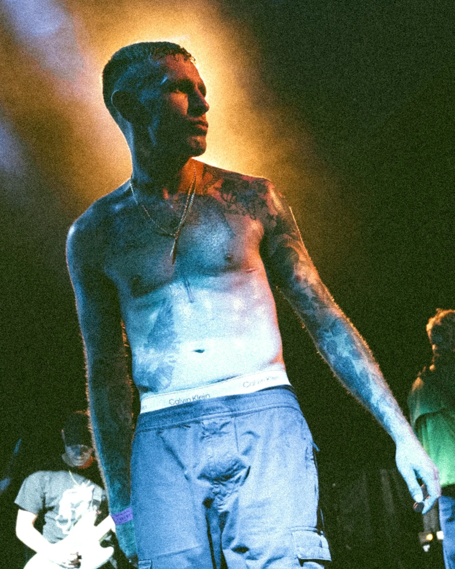 A shirtless man with tattoos and short hair stands in a dimly lit environment with a golden glow behind him. In the background, there are other people, one playing a guitar.