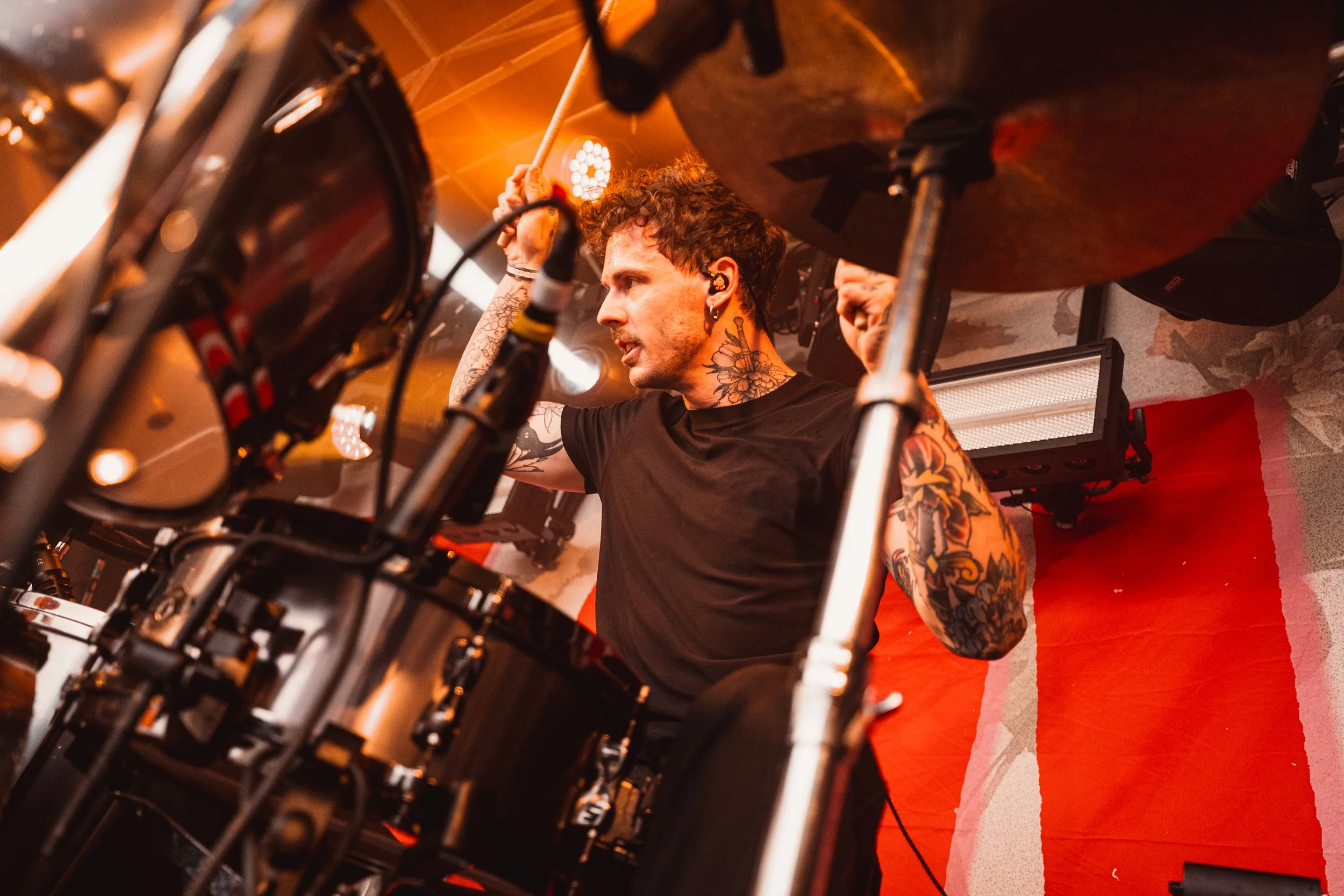 A male drummer with tattoos playing drums on stage, looking intensely at his instrument. Stage lighting with orange hues reflects off the drums.