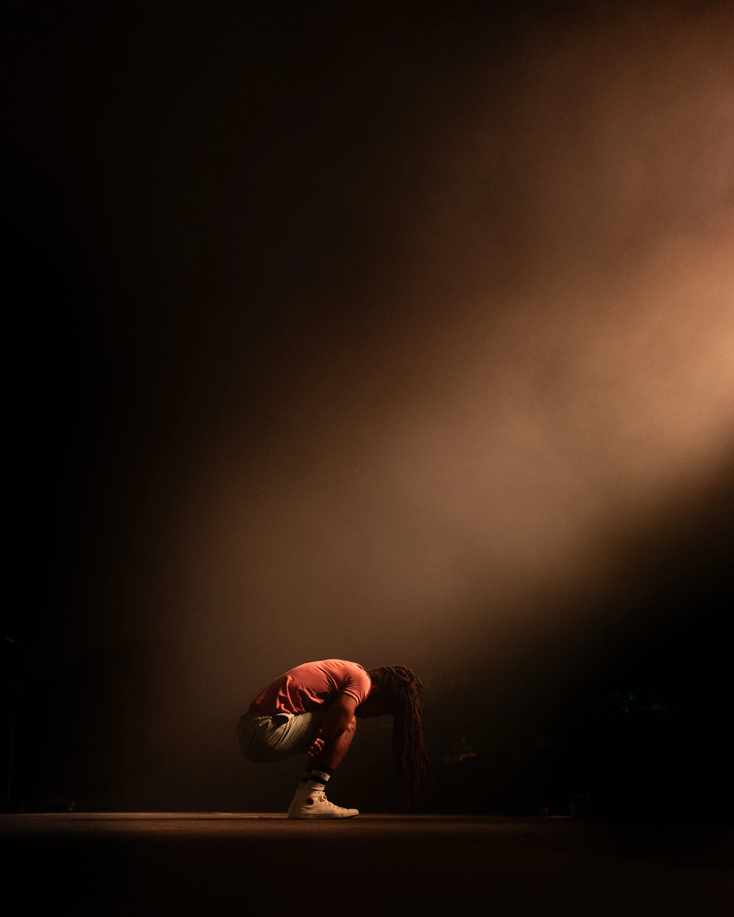 A person with long dreadlocks crouches down on stage in a dimly lit environment with a hazy, colorful background.