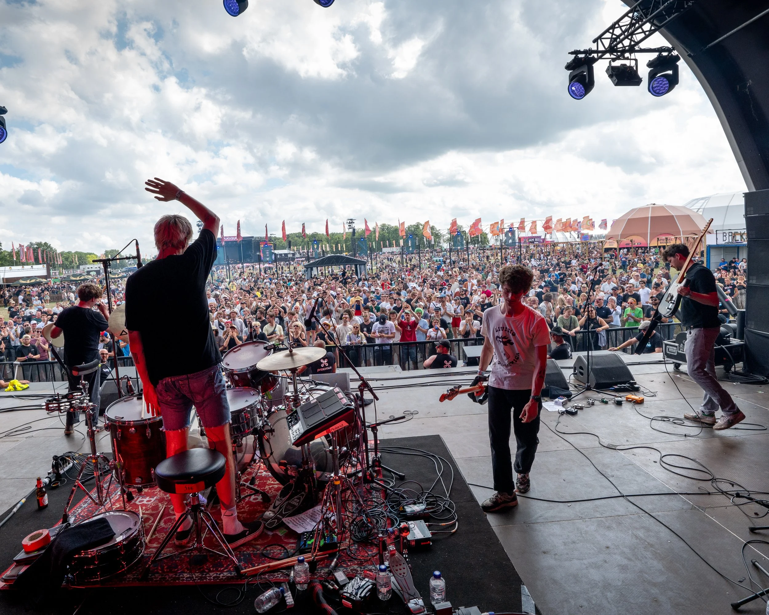 View of a live outdoor concert stage with band members and a large audience under cloudy sky.