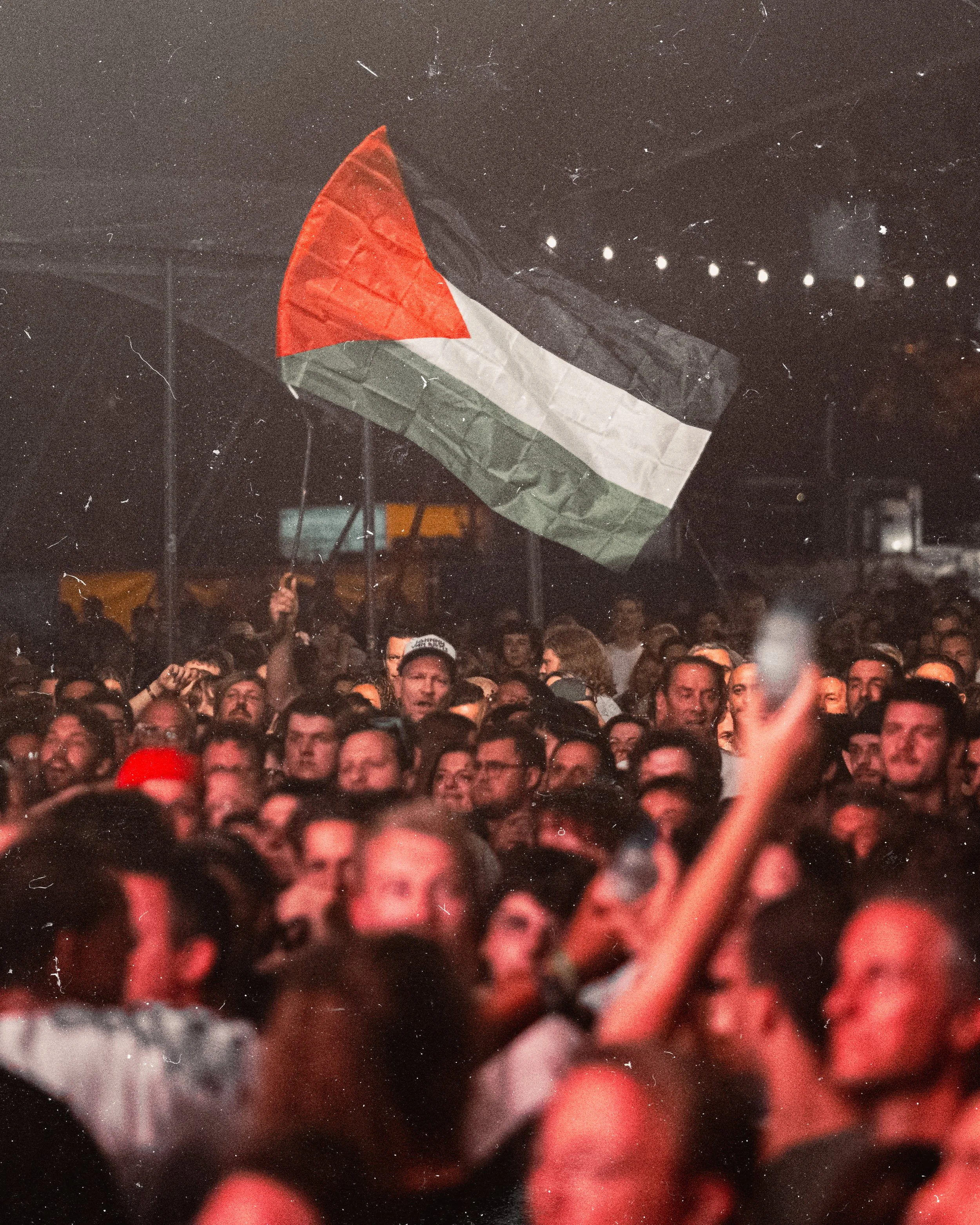 A crowd of people gathered at a nighttime event, with an individual holding a Palestinian flag.