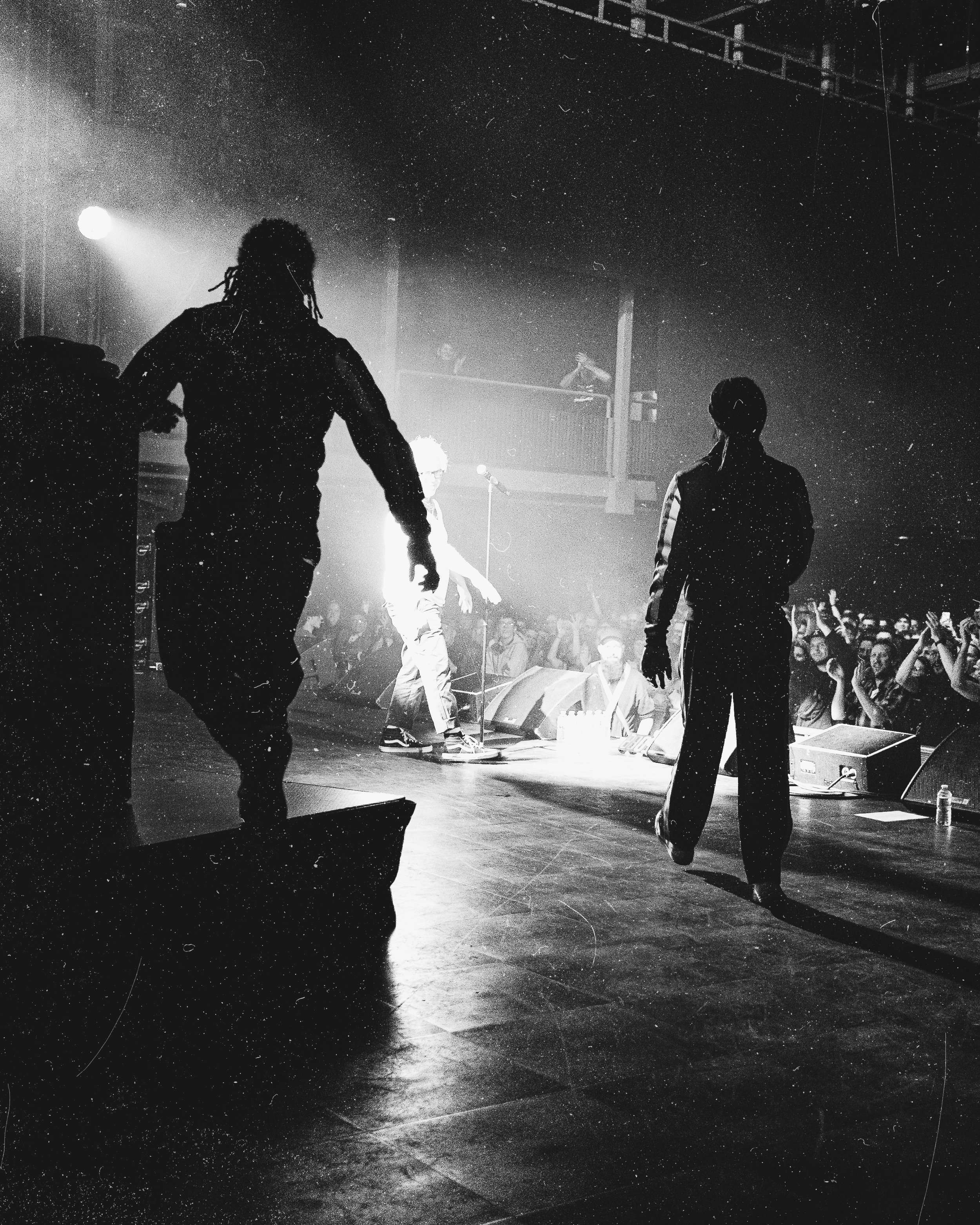Black and white photo of a music concert with two silhouette figures on stage and a crowd in front.