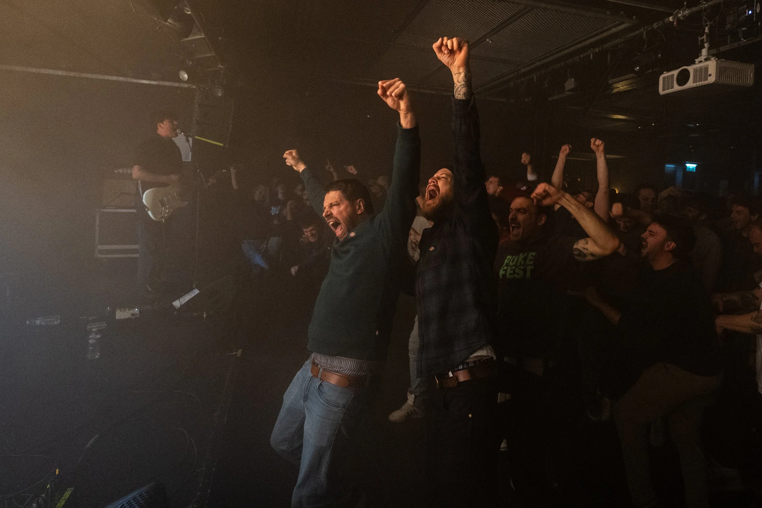 Crowd at a concert or event cheering with hands raised, some with fists in the air, in a dimly lit venue.