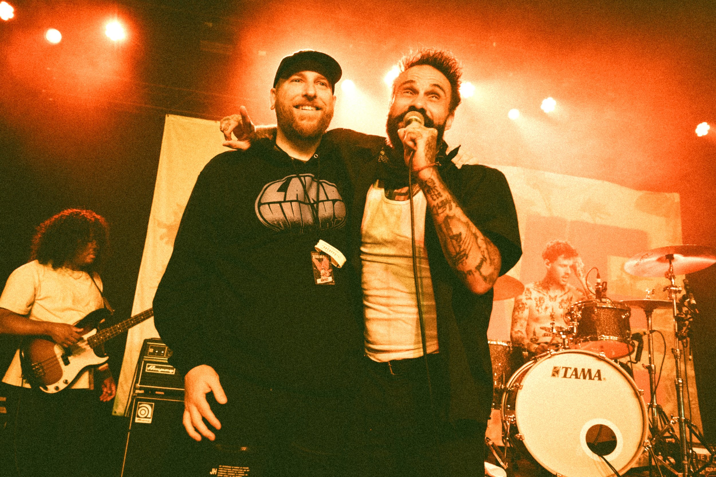 Two men standing closely on stage, one holding a microphone, with a band performing in the background under orange stage lights.