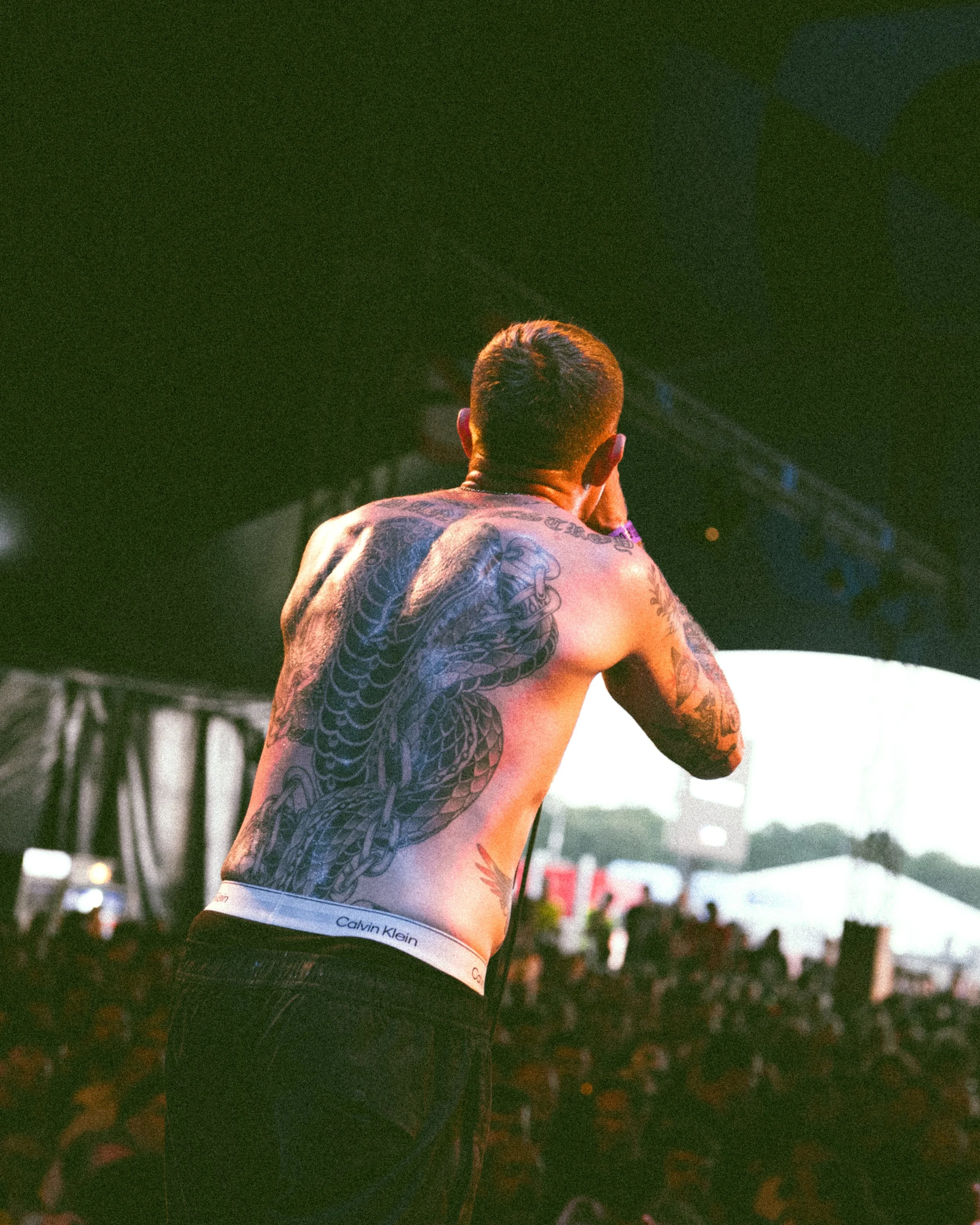 A shirtless male performer with tattoos on his back and arms, seen from behind, speaking into a microphone on stage at an outdoor concert.