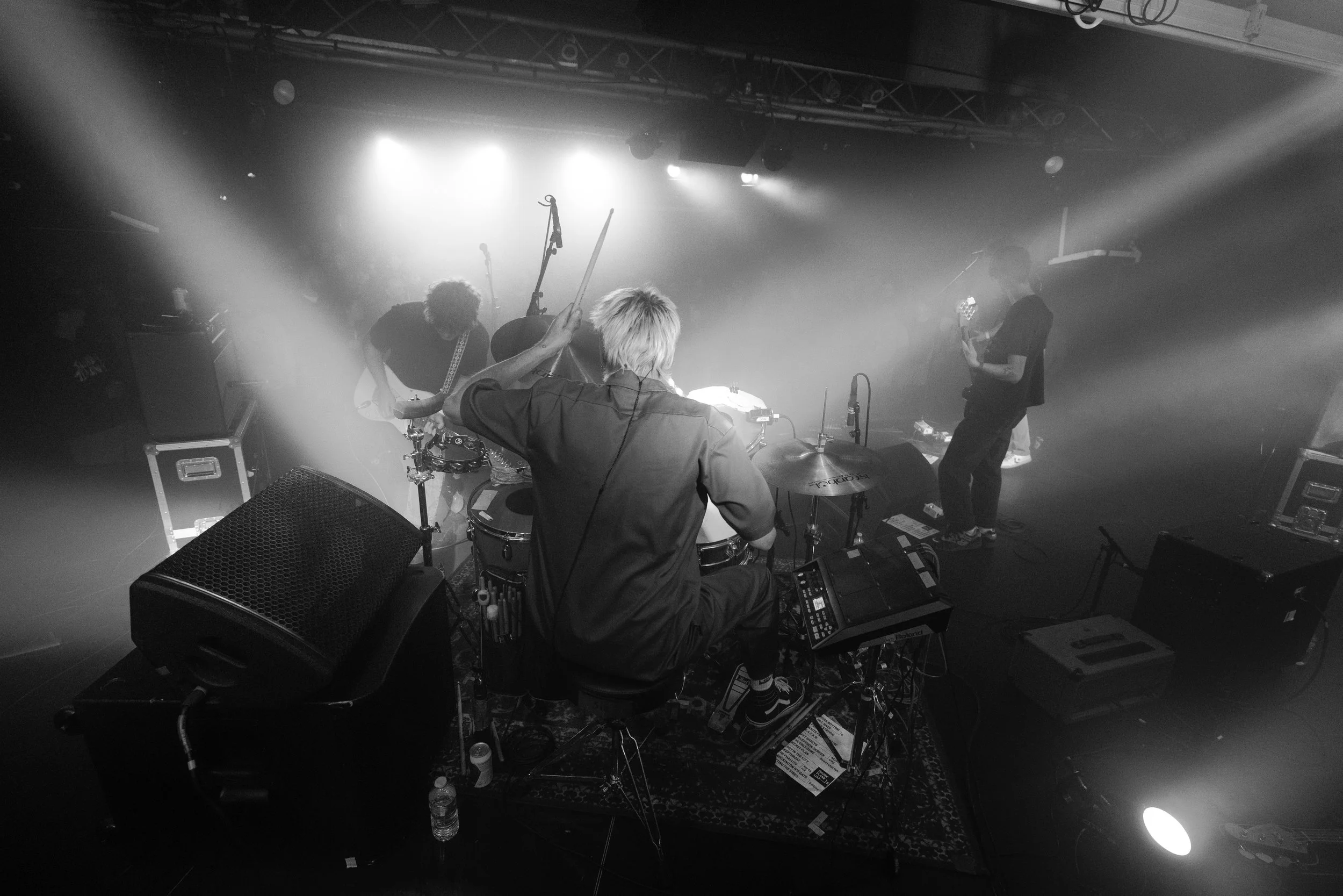 Black and white photo of a band performing on stage with a drummer in the foreground, other band members playing guitars. Stage lighting creates a hazy atmosphere.