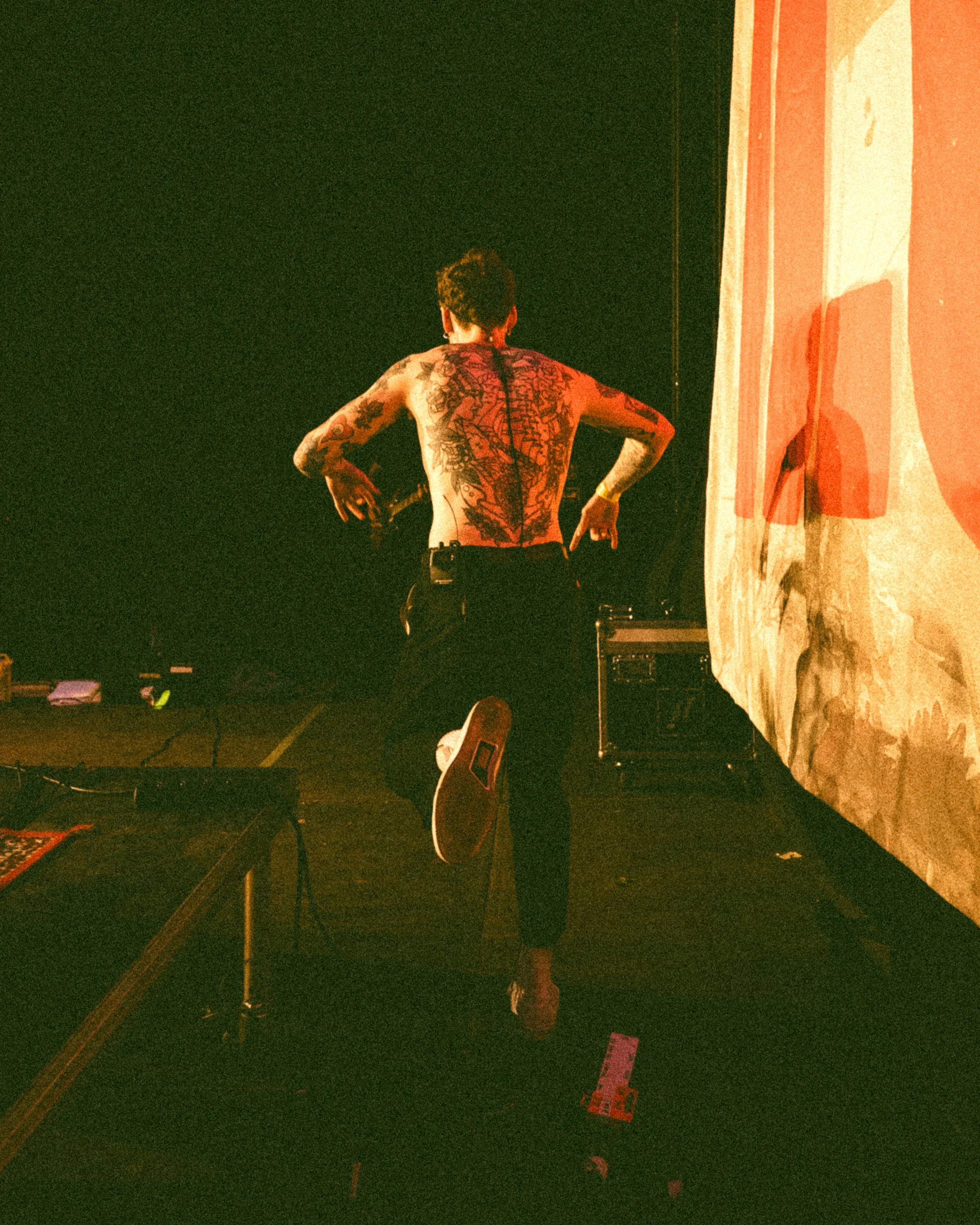 A shirtless man with tattoos on his back is standing on one leg with his arms bent, facing away from the camera, in a dimly lit room with a large projection screen to the side.