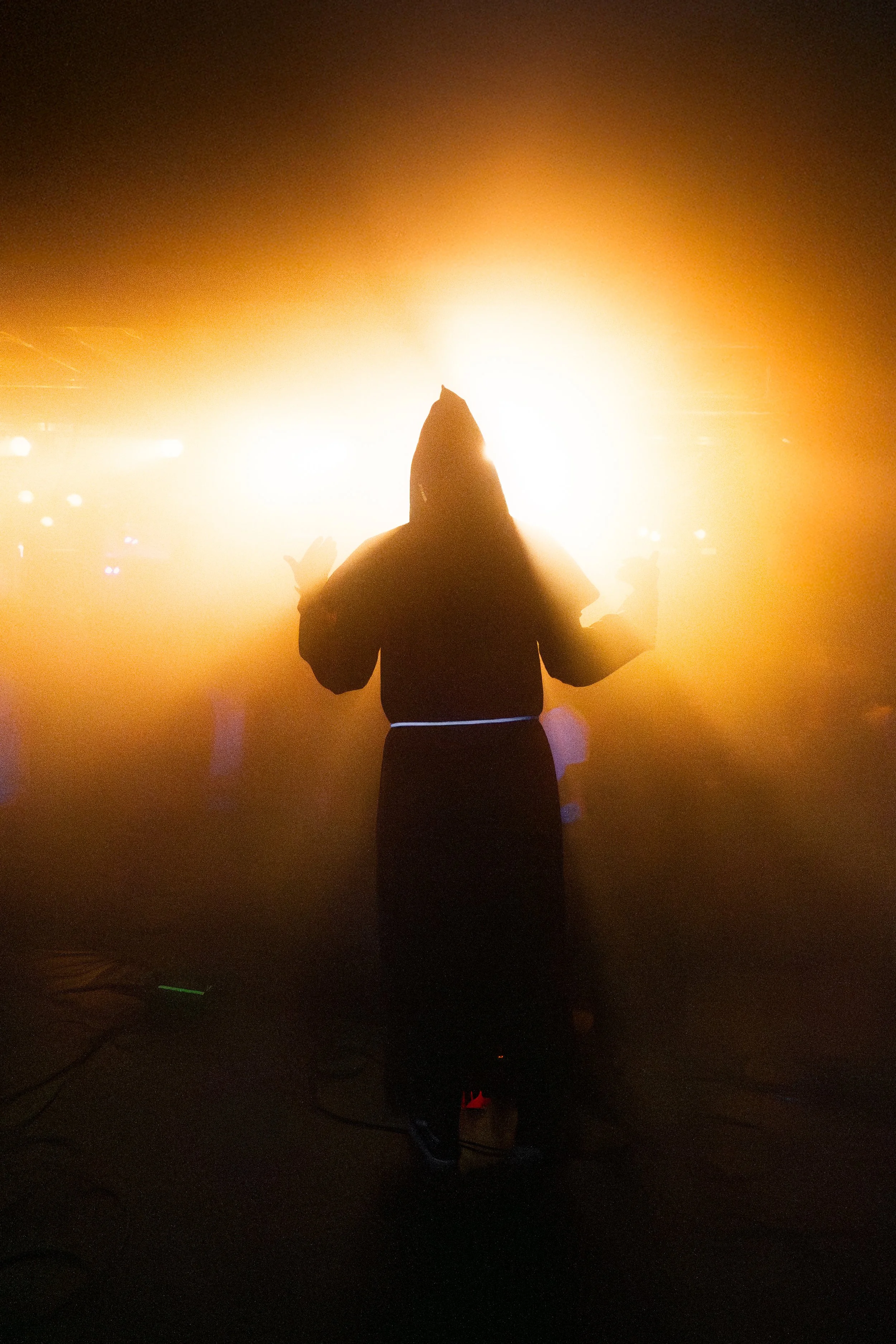 Silhouette of a person standing with arms raised, backlit by bright orange and white lights in a dark setting, possibly a concert or nightclub.