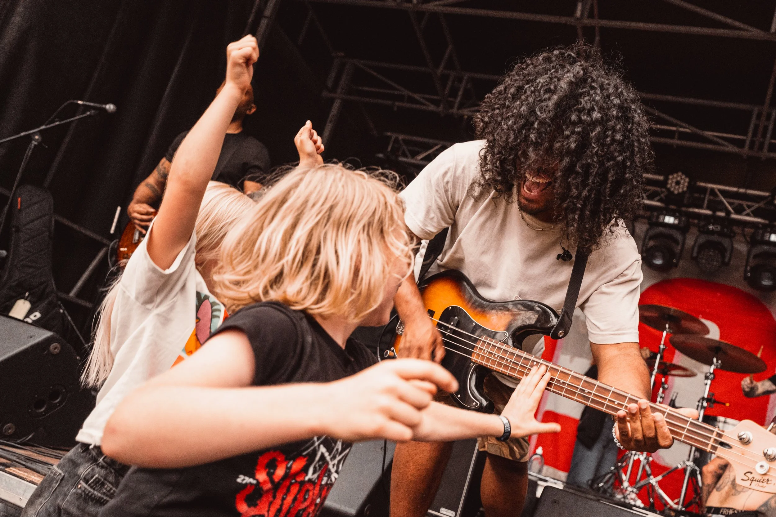 A man with curly hair plays bass guitar on stage while two young girls, one with blonde hair and one with fair hair, sing and dance along. The stage has musical equipment and a drummer in the background.