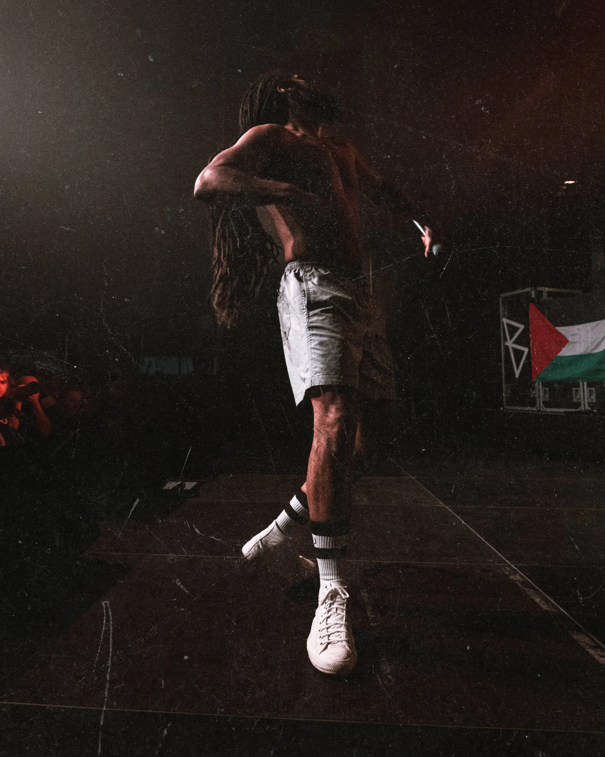 A person with dreadlocks standing on stage, holding a microphone, with a Palestinian flag displayed in the background.