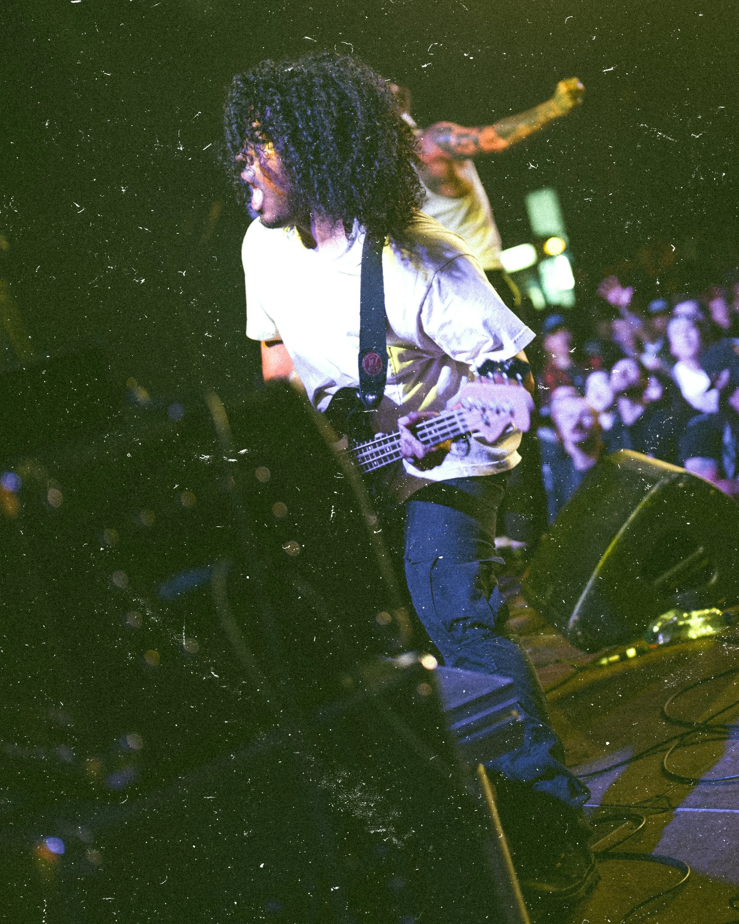 A musician with curly hair passionately plays an electric guitar on stage in front of a crowd at a concert.