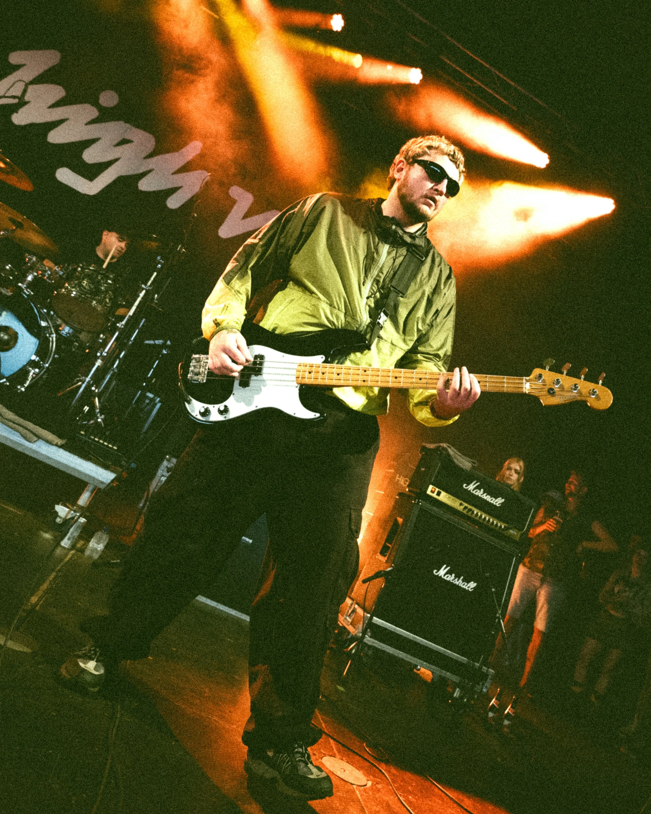 A musician wearing sunglasses and a green windbreaker playing a black and white bass guitar on stage with orange lighting, backed by a drum set and Marshall amplifiers.