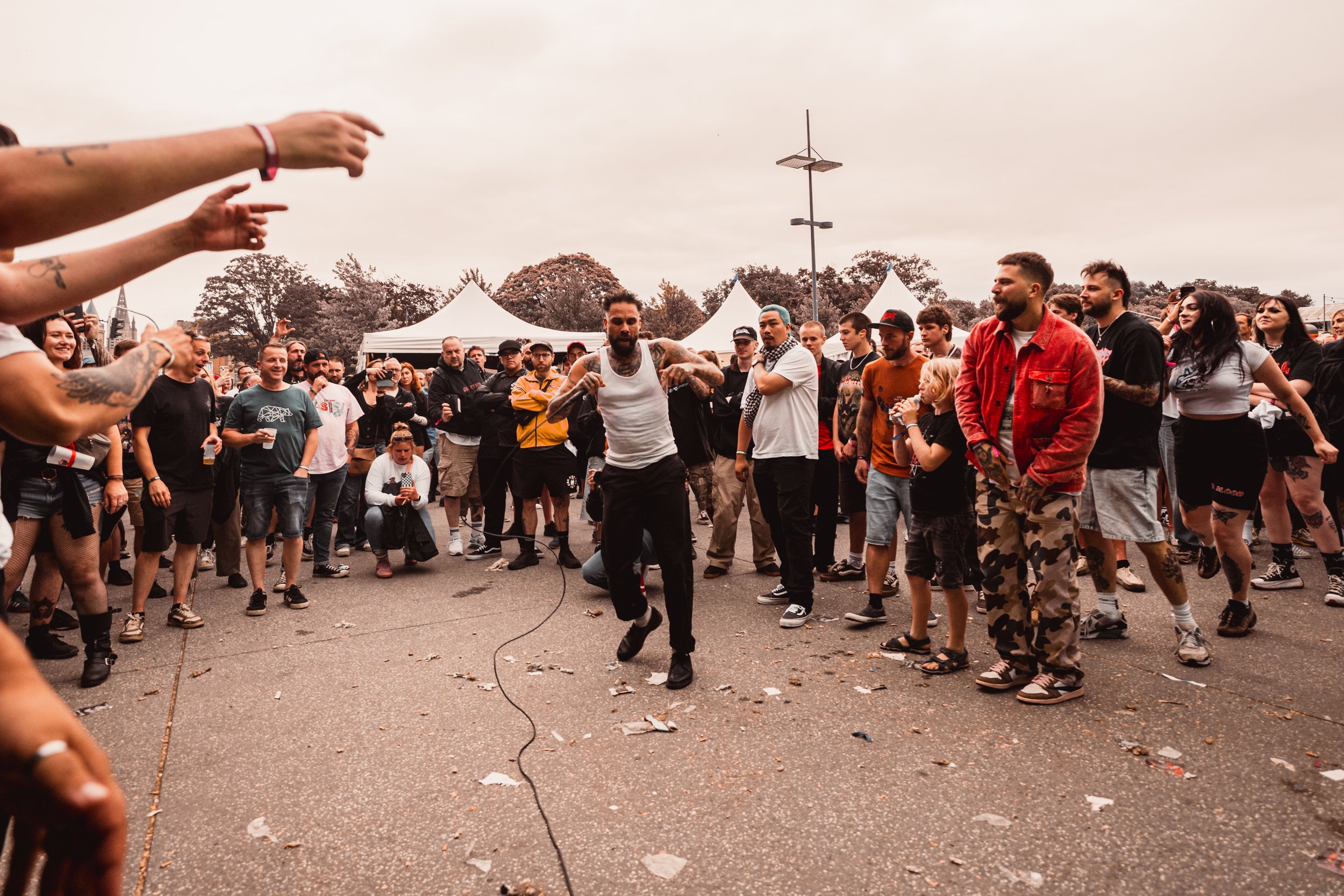 A crowd of people attending an outdoor concert or festival on a cloudy day, with a man performing or dancing in the center, wearing a white tank top and black pants, surrounded by others standing and observing.