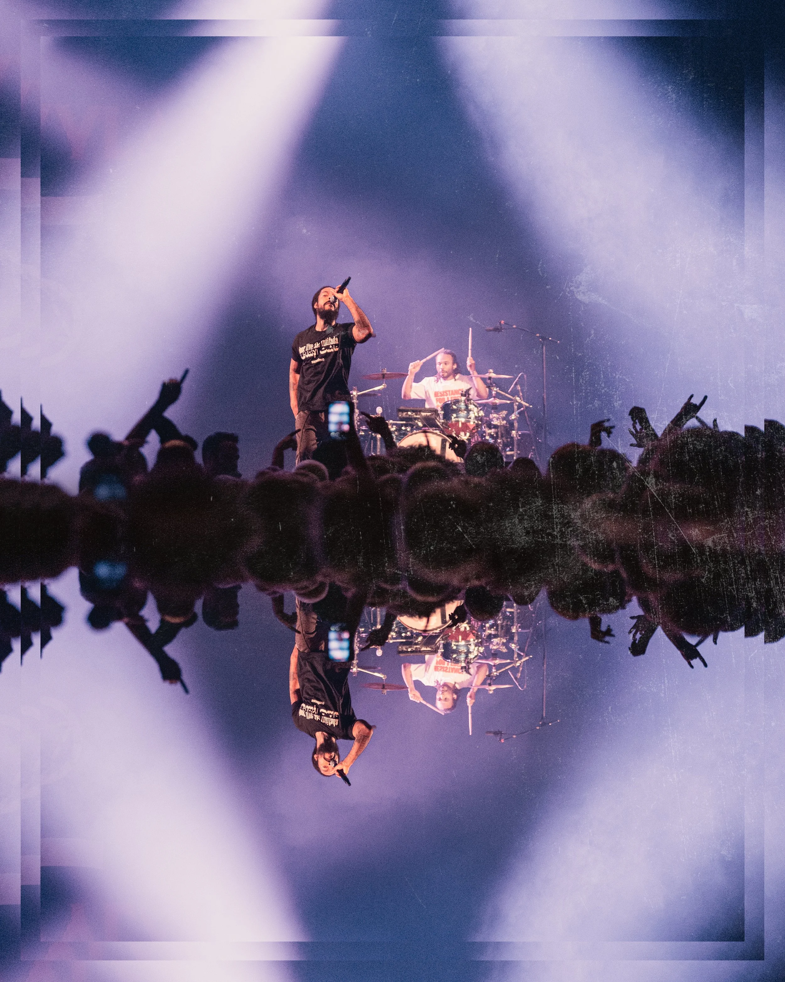 A live concert with a vocalist singing into a microphone and a drummer playing behind him, with audience members in the foreground and stage lights creating a dramatic atmosphere.