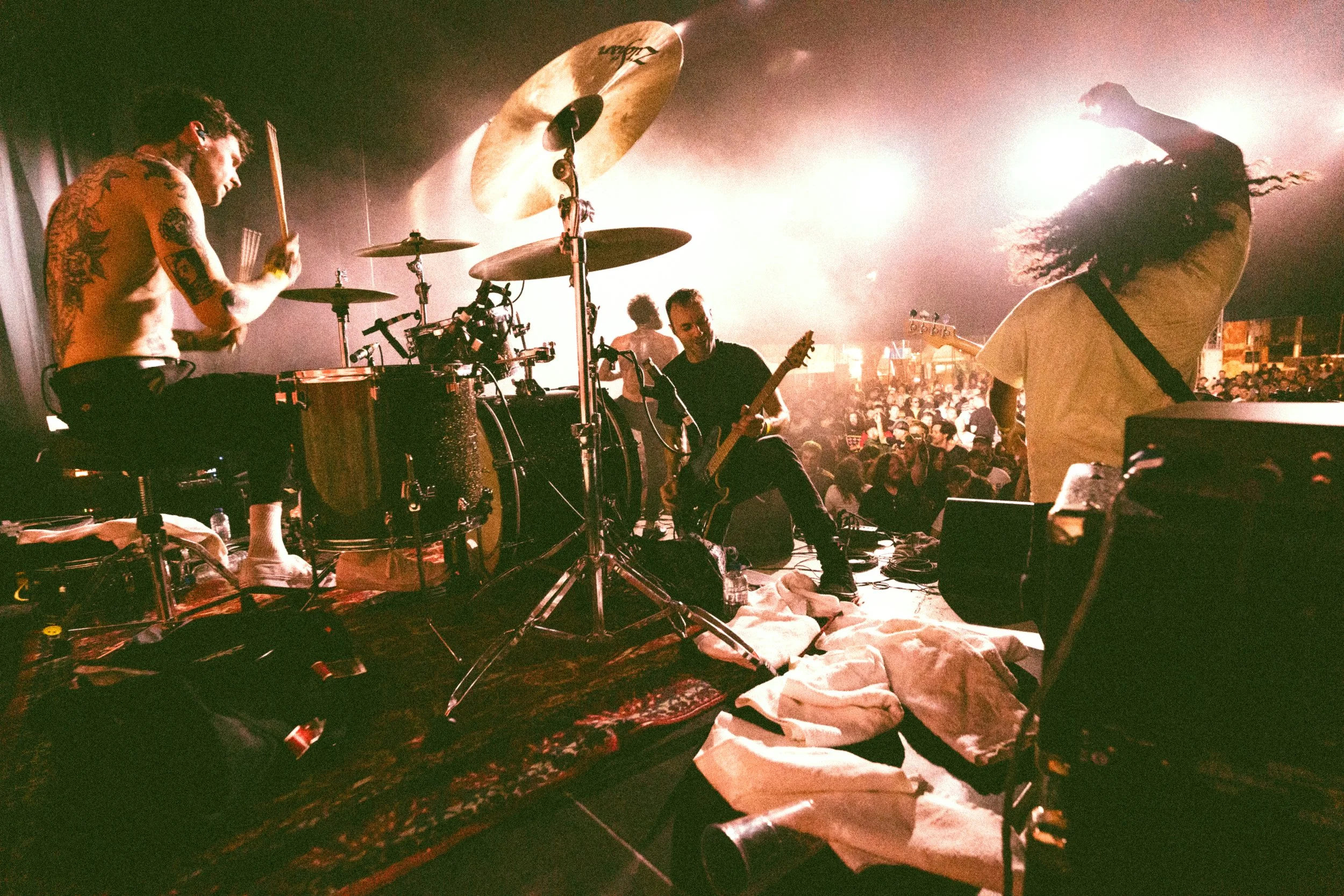 Band performing live on stage with drums, guitar, and bass in front of a crowd at concert venue, with bright lighting and audience visible in the background.