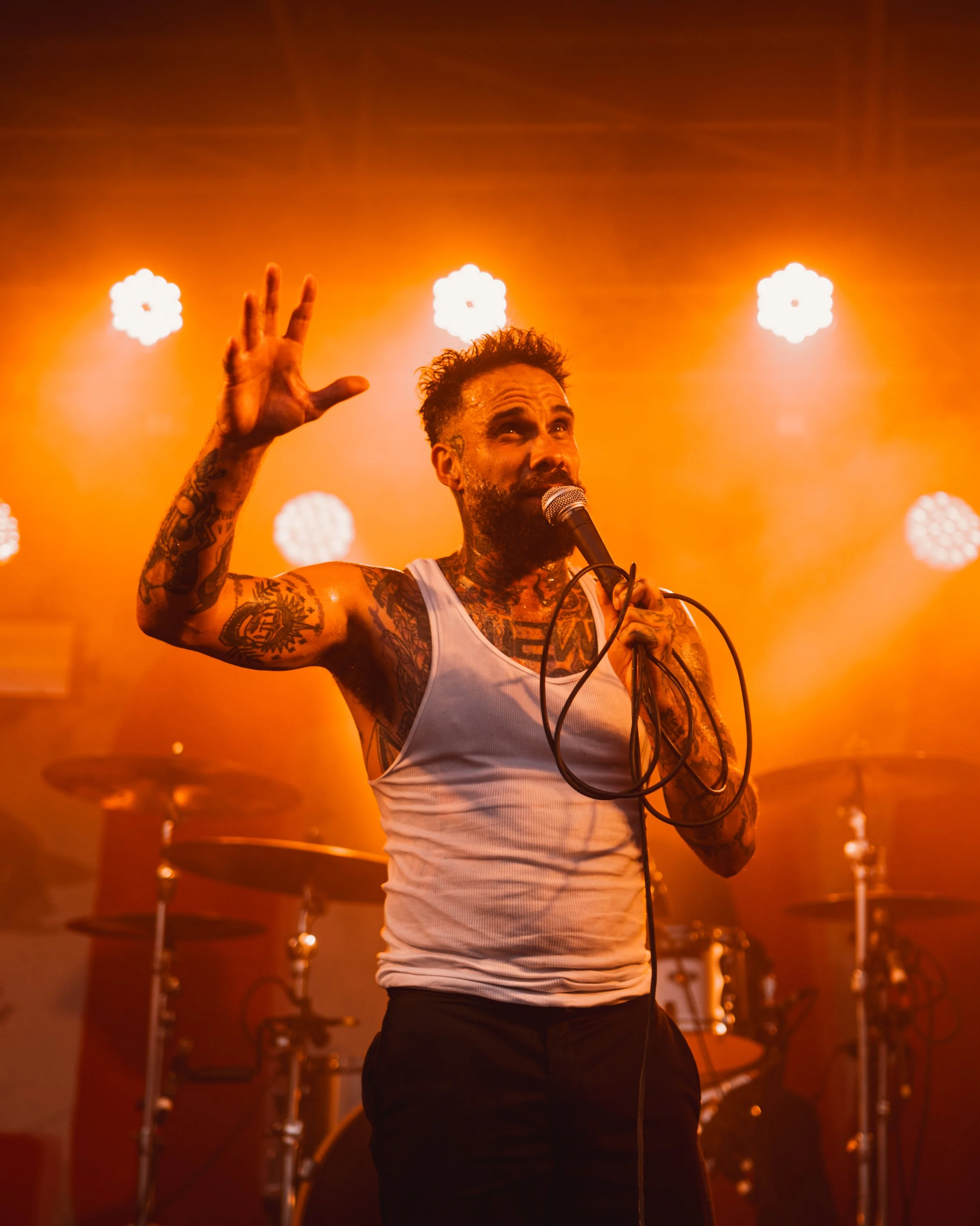 Tattooed male singer in a white tank top holding a microphone, raising his hand on stage with orange lighting and a drum set in the background.