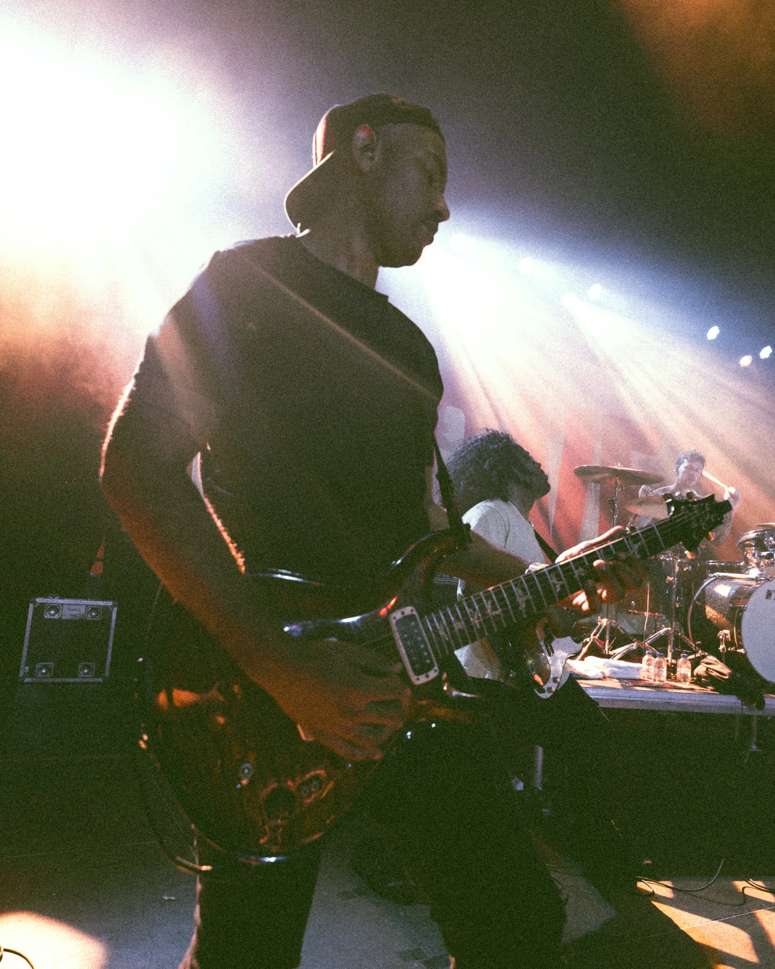 Silhouetted male guitarist performing on stage with colorful stage lights, other band members playing instruments in the background.