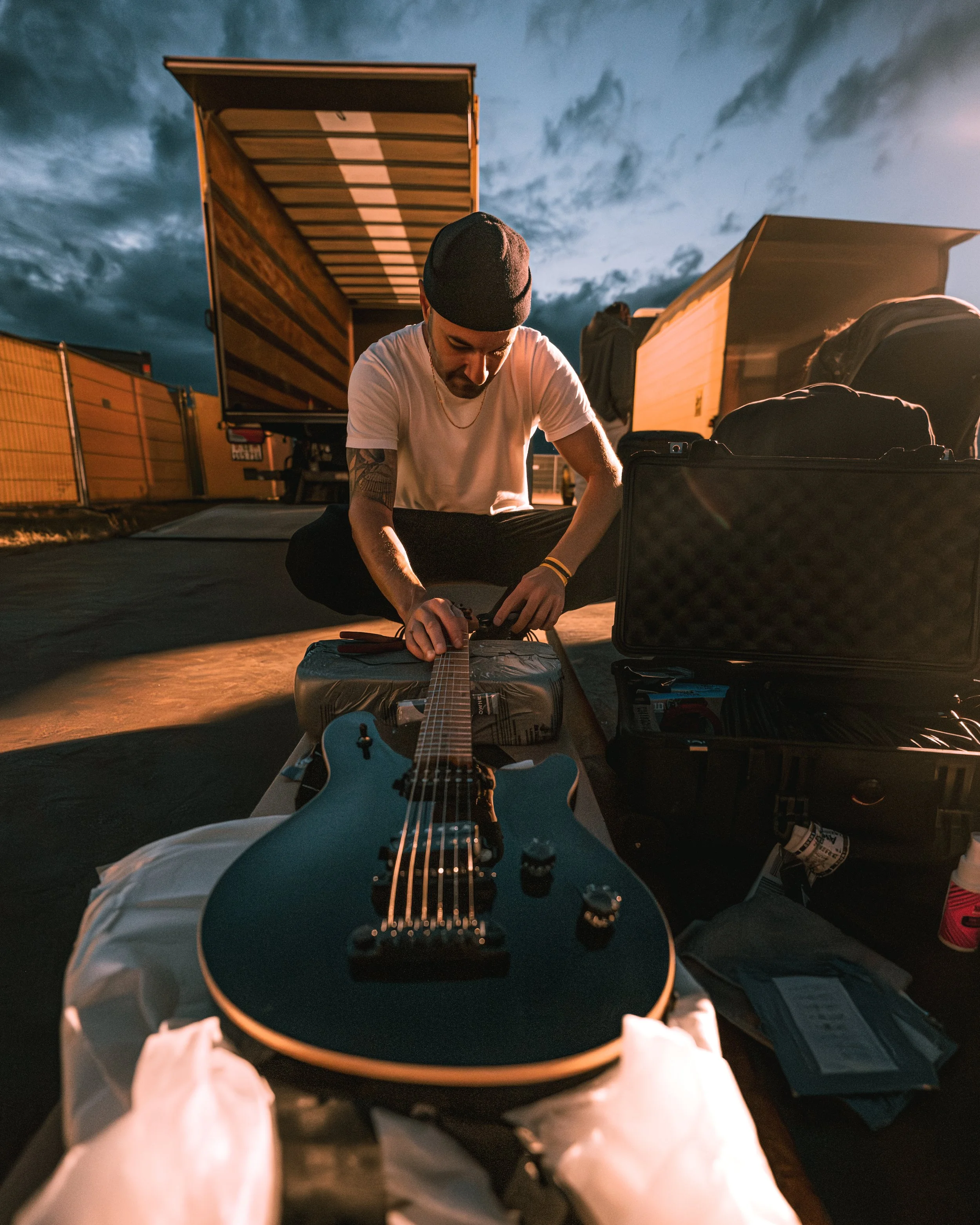 A man sitting on the ground, packing a guitar case opened in front of him, with trucks and a fence in the background during sunset or dusk.