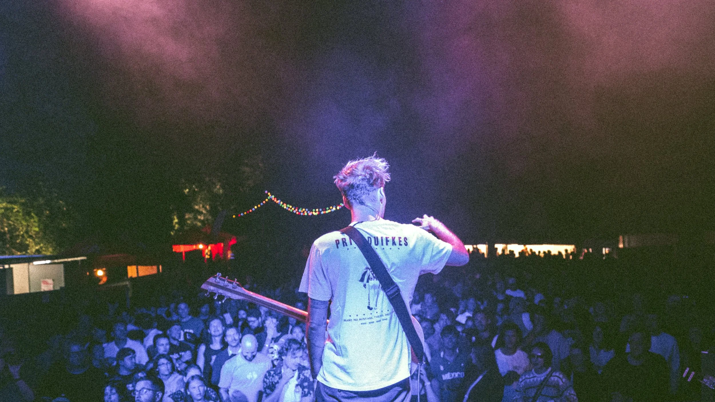 Performer on stage with guitar facing a large audience in a dark, colorful atmosphere with string lights in the background.