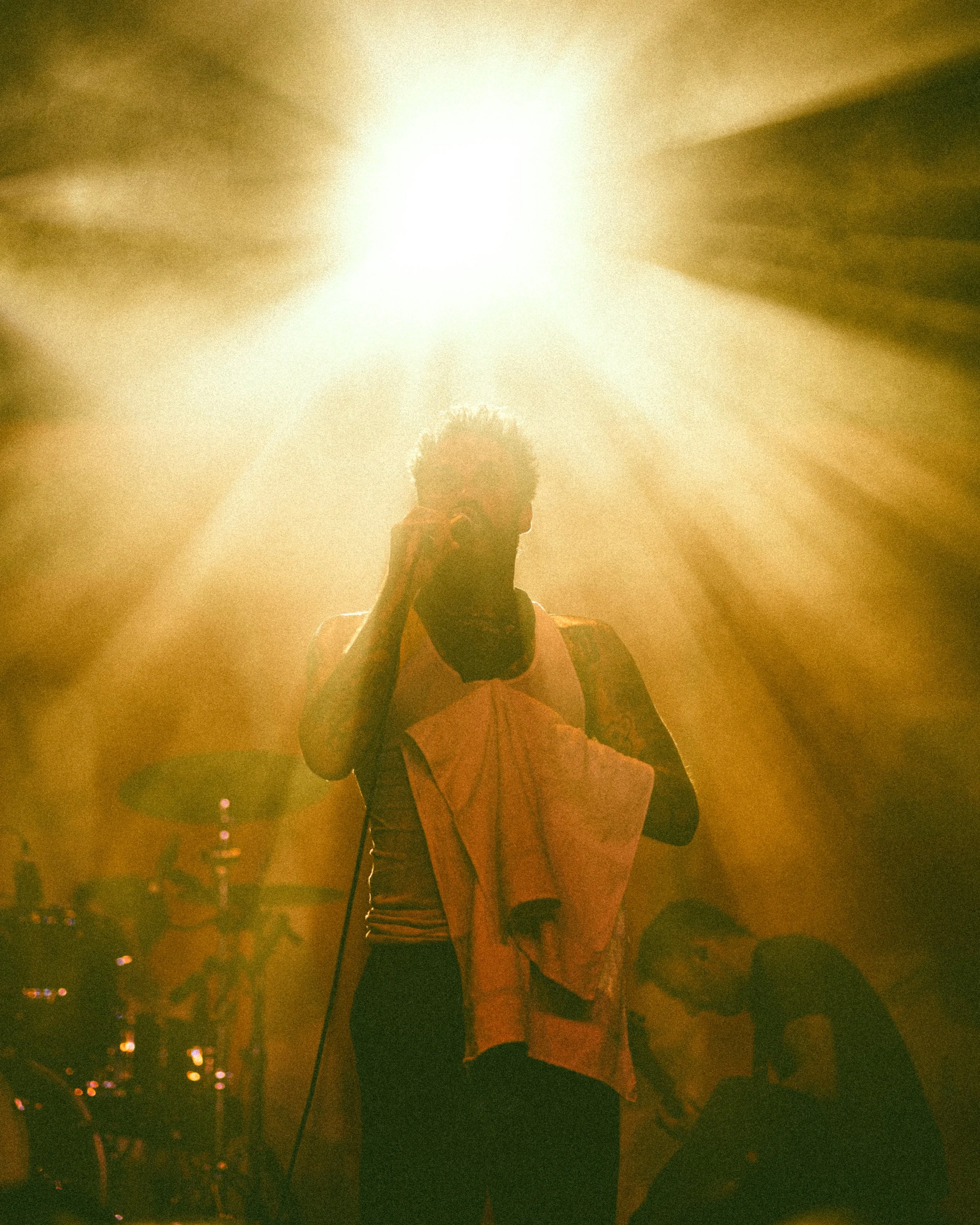 A person holding a microphone and a towel, on stage with bright, golden stage lighting, and a band setup in the background.