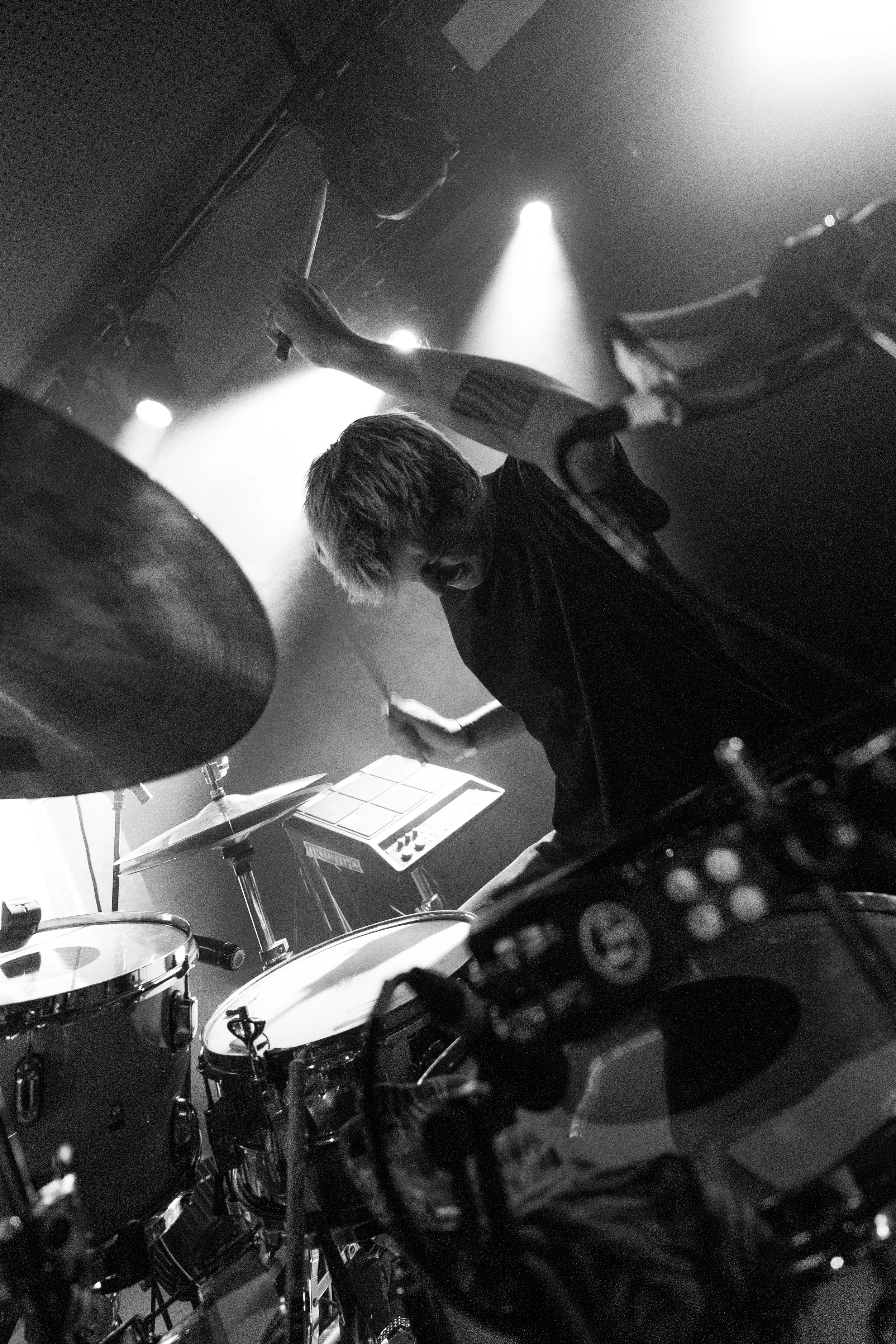Black and white photo of a drummer performing on stage with drums and electronic music equipment, illuminated by stage lights.