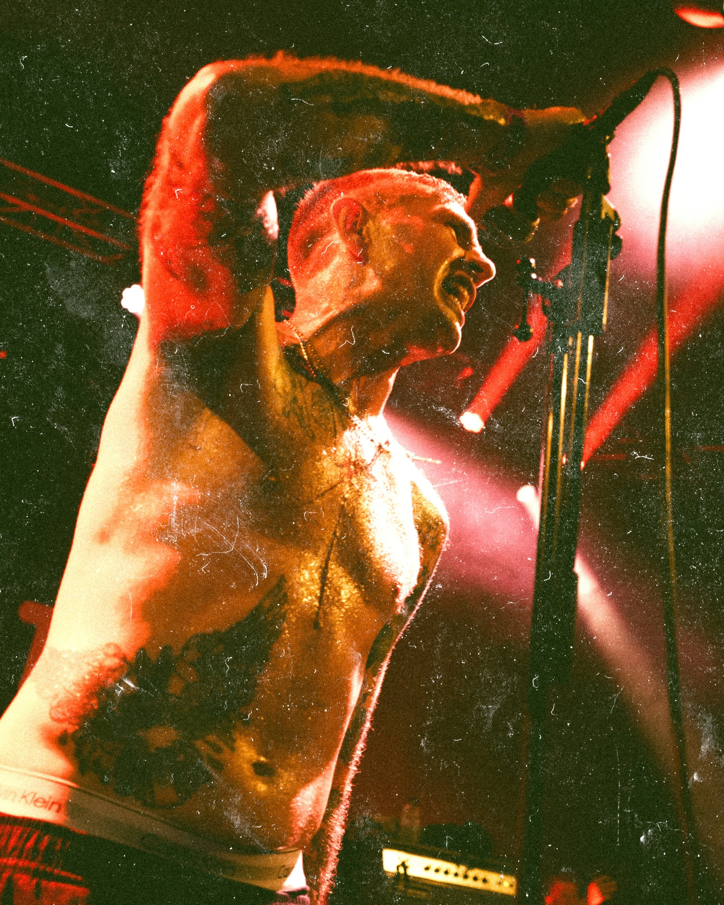 A shirtless male performer singing passionately on stage with intense lighting and visible body tattoos.