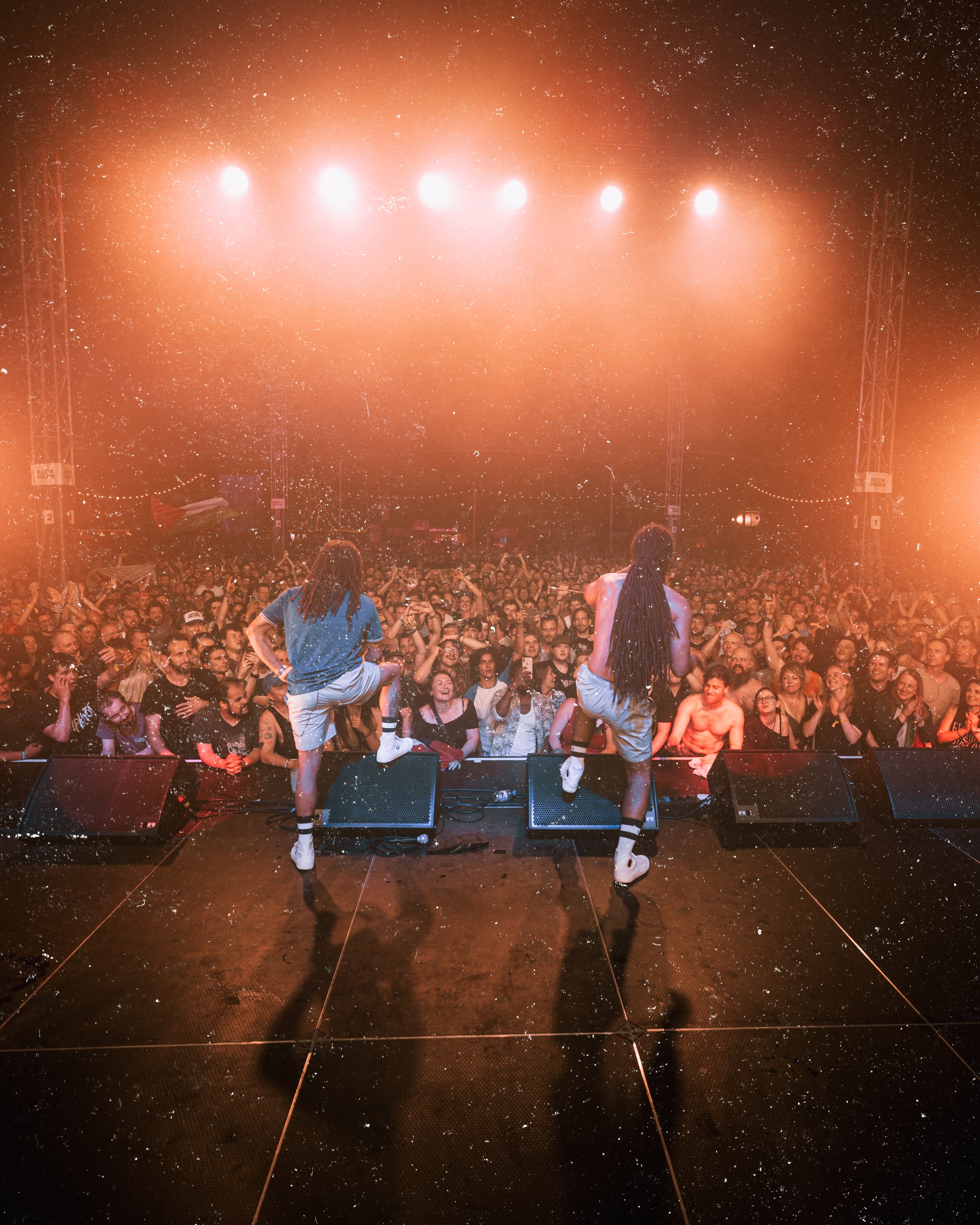 Two performers on stage with dreadlocks, facing a large crowd at a concert, under bright orange lighting and a starry background.