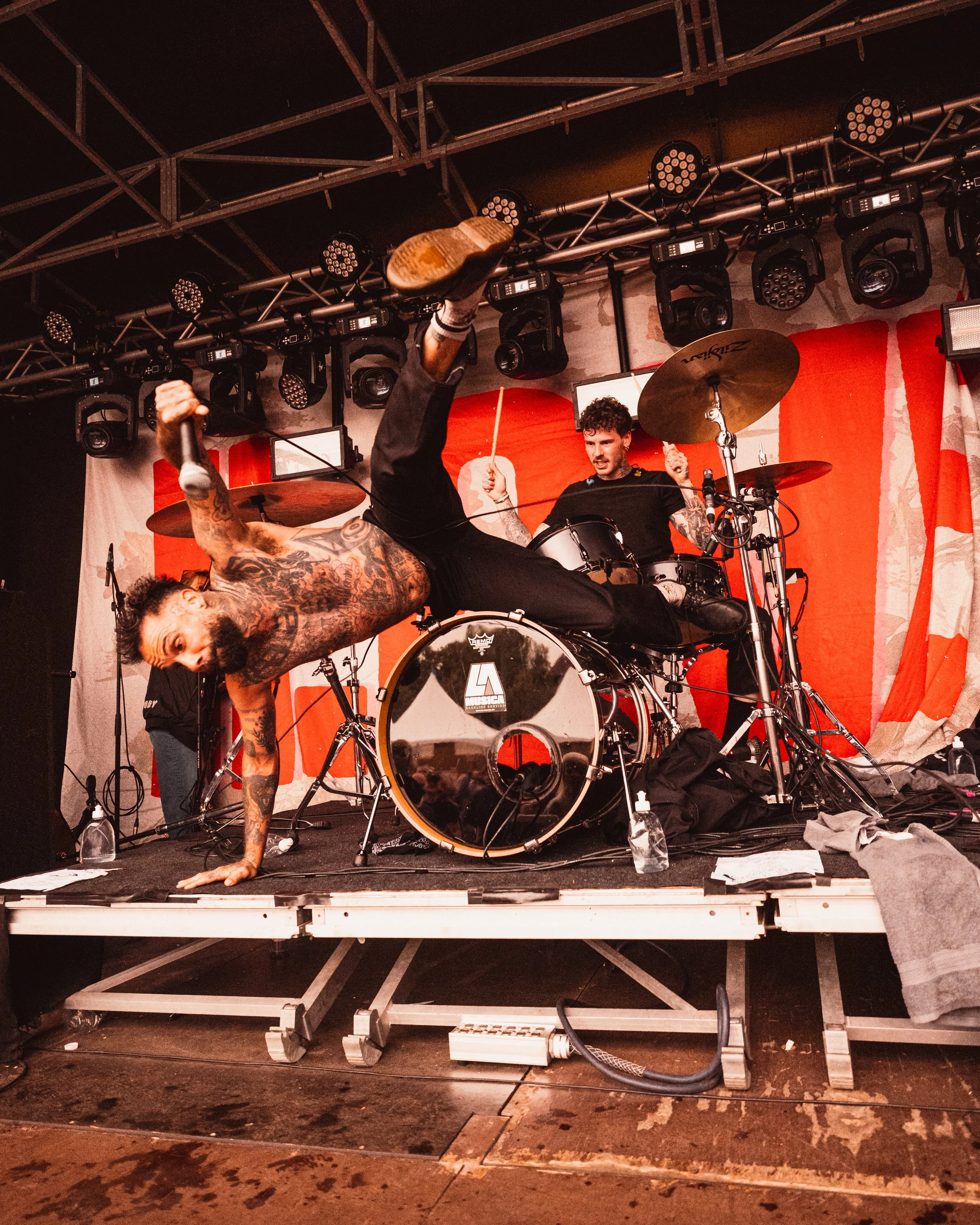 A singer performs on stage while doing a handstand, and a drummer plays behind him. The stage has a red backdrop with a large white design, and various stage lights are overhead.