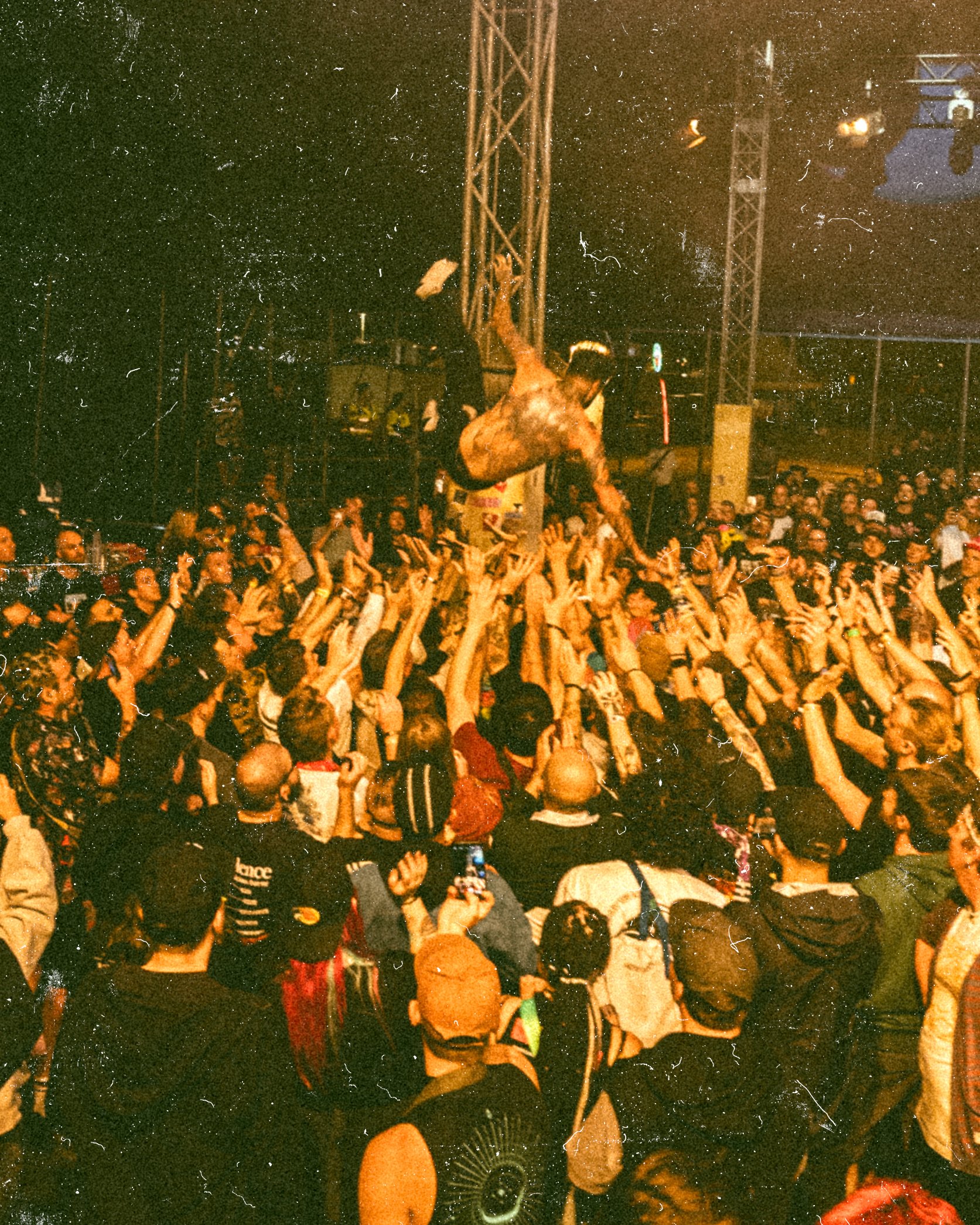 A large crowd at a concert or festival with a person tossing an object into the air above the audience, illuminated by stage lights.