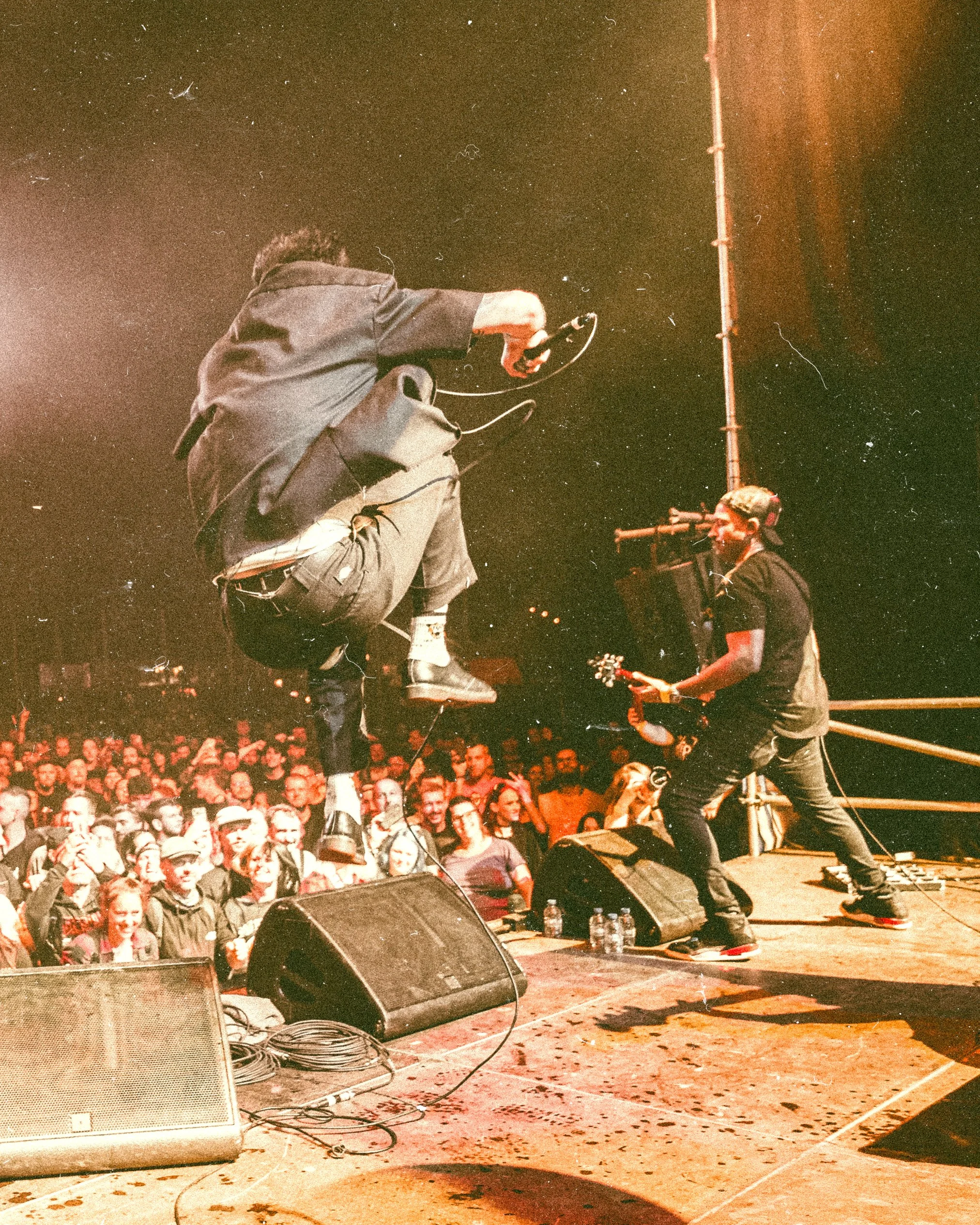 A person is mid-air on stage during a concert, jumping with a microphone, while a guitarist plays nearby. An audience is watching and taking photos in front of the stage.