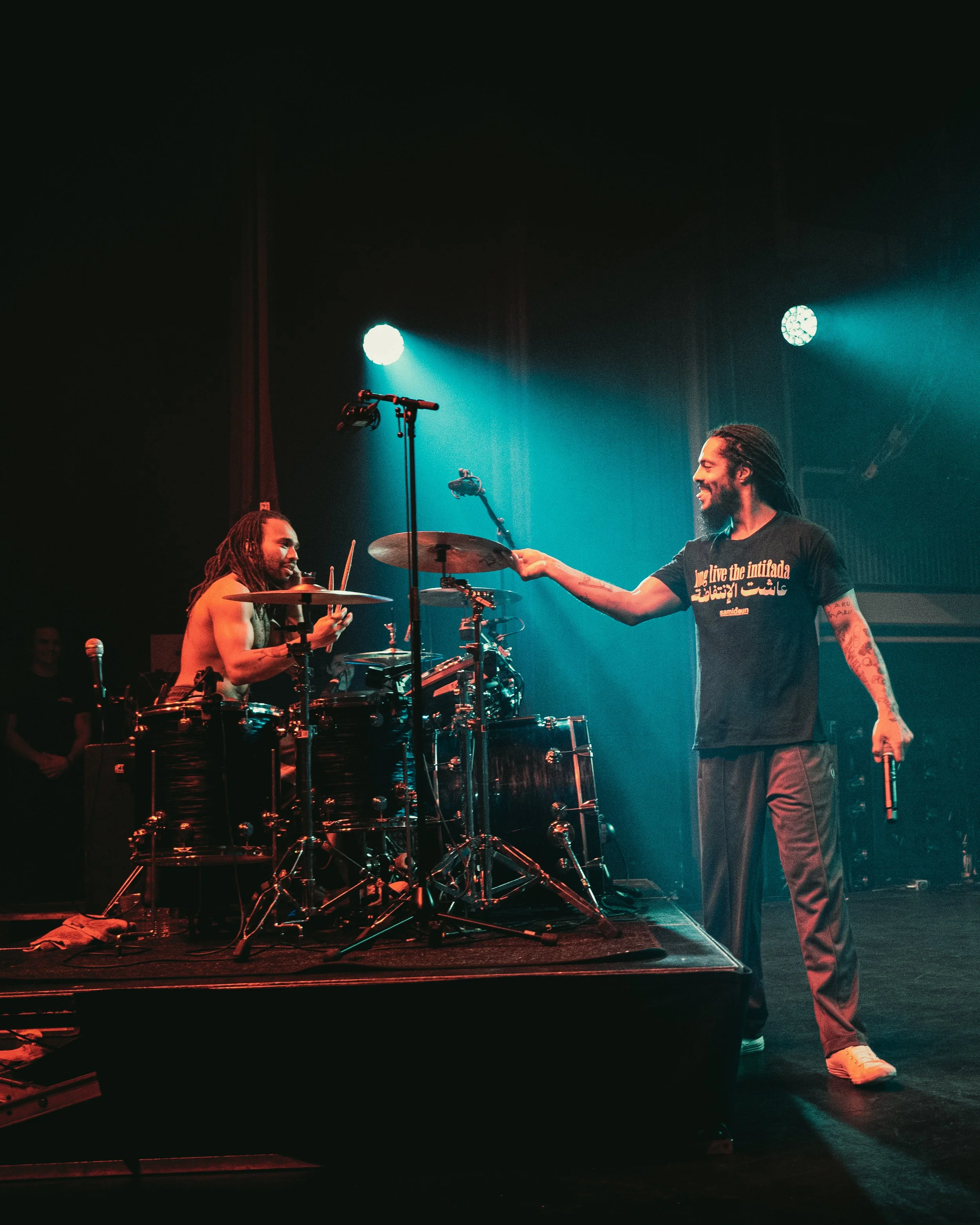 Two musicians performing on stage under blue and teal lights. One is playing drums, and the other is with a microphone in one hand and reaching out to the drummer.