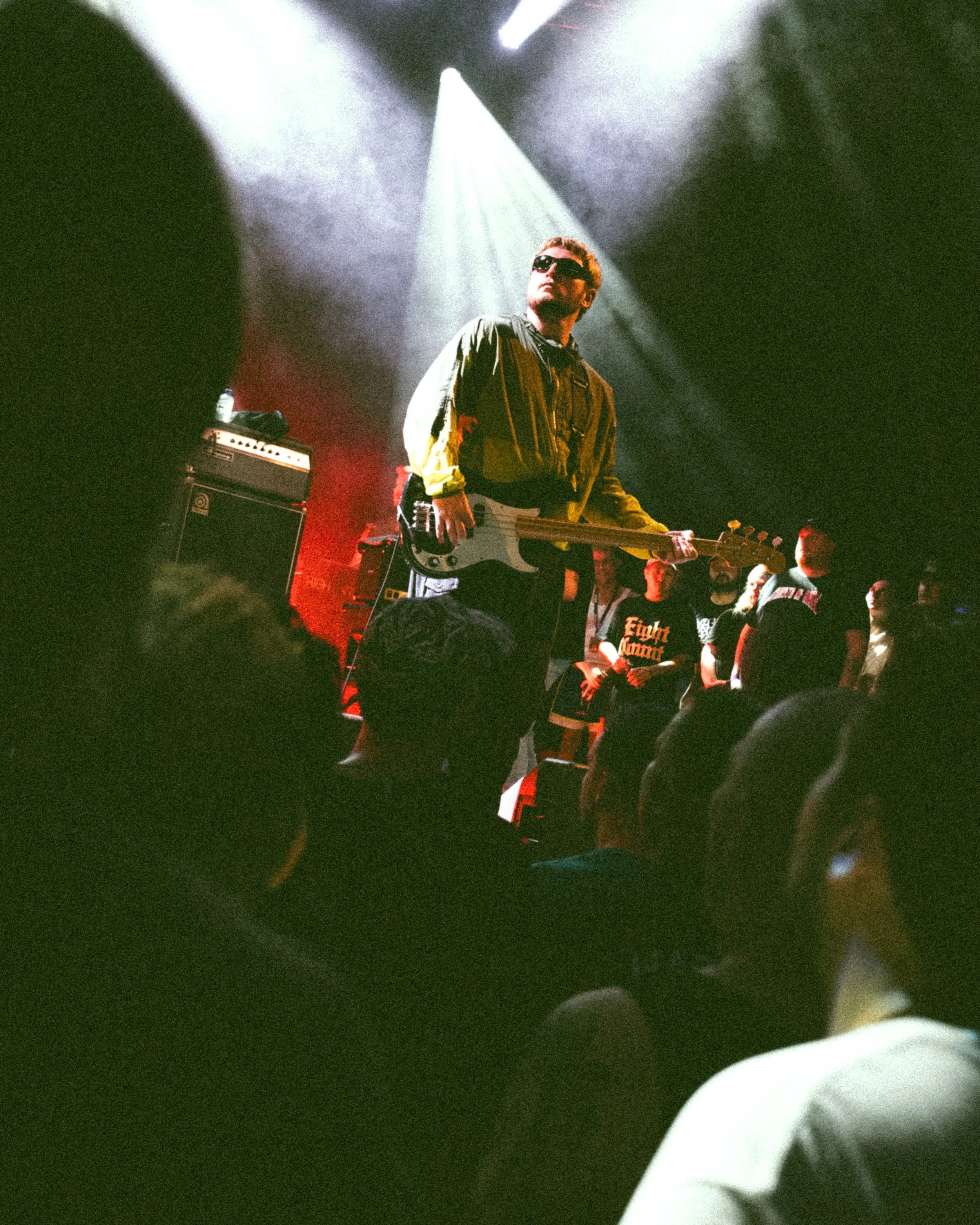 A man wearing sunglasses and a green jacket playing an electric guitar on stage during a concert, with a crowd watching in the foreground.