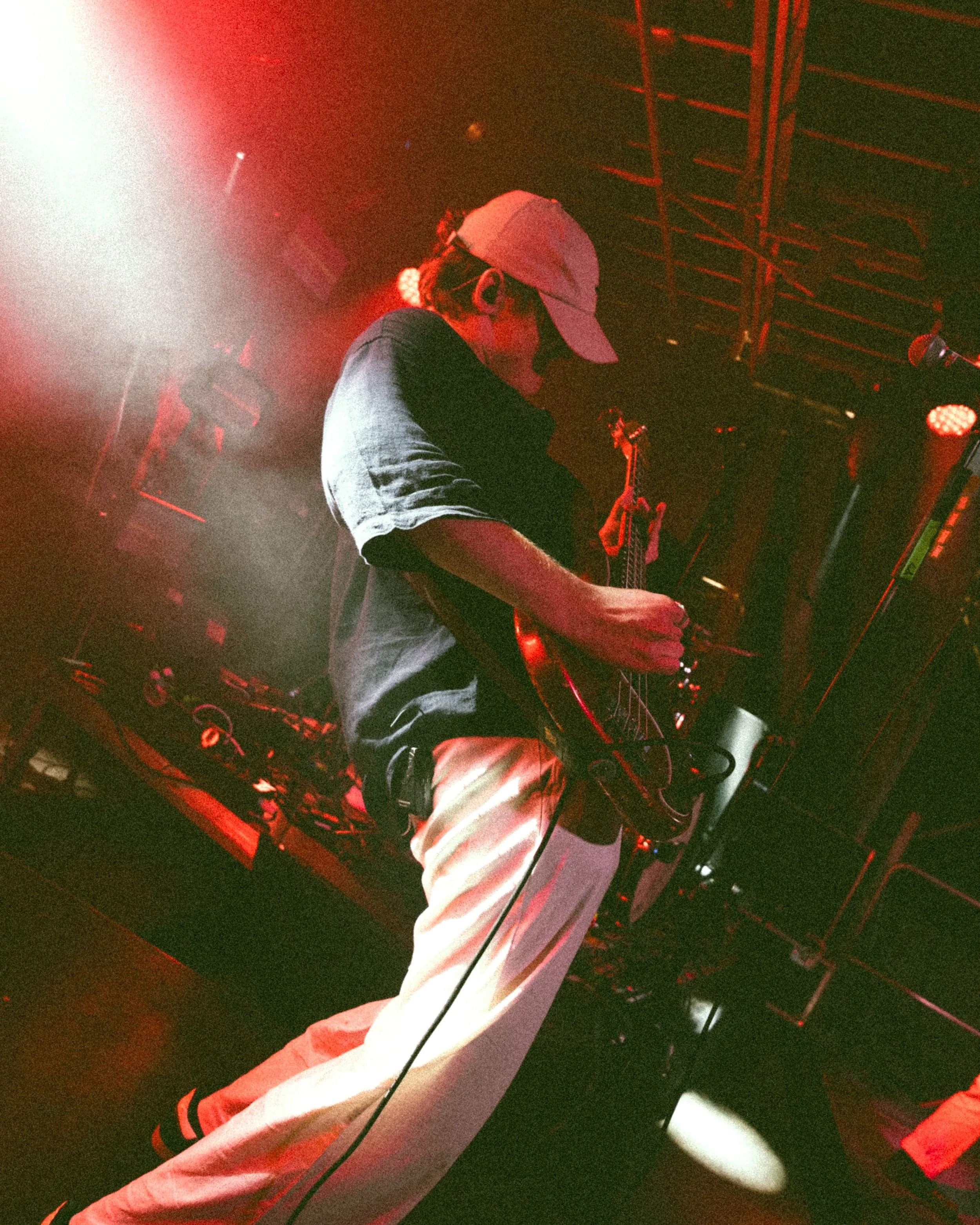 A person wearing a baseball cap, dark shirt, and light-colored pants is playing an electric guitar on stage with red lighting.