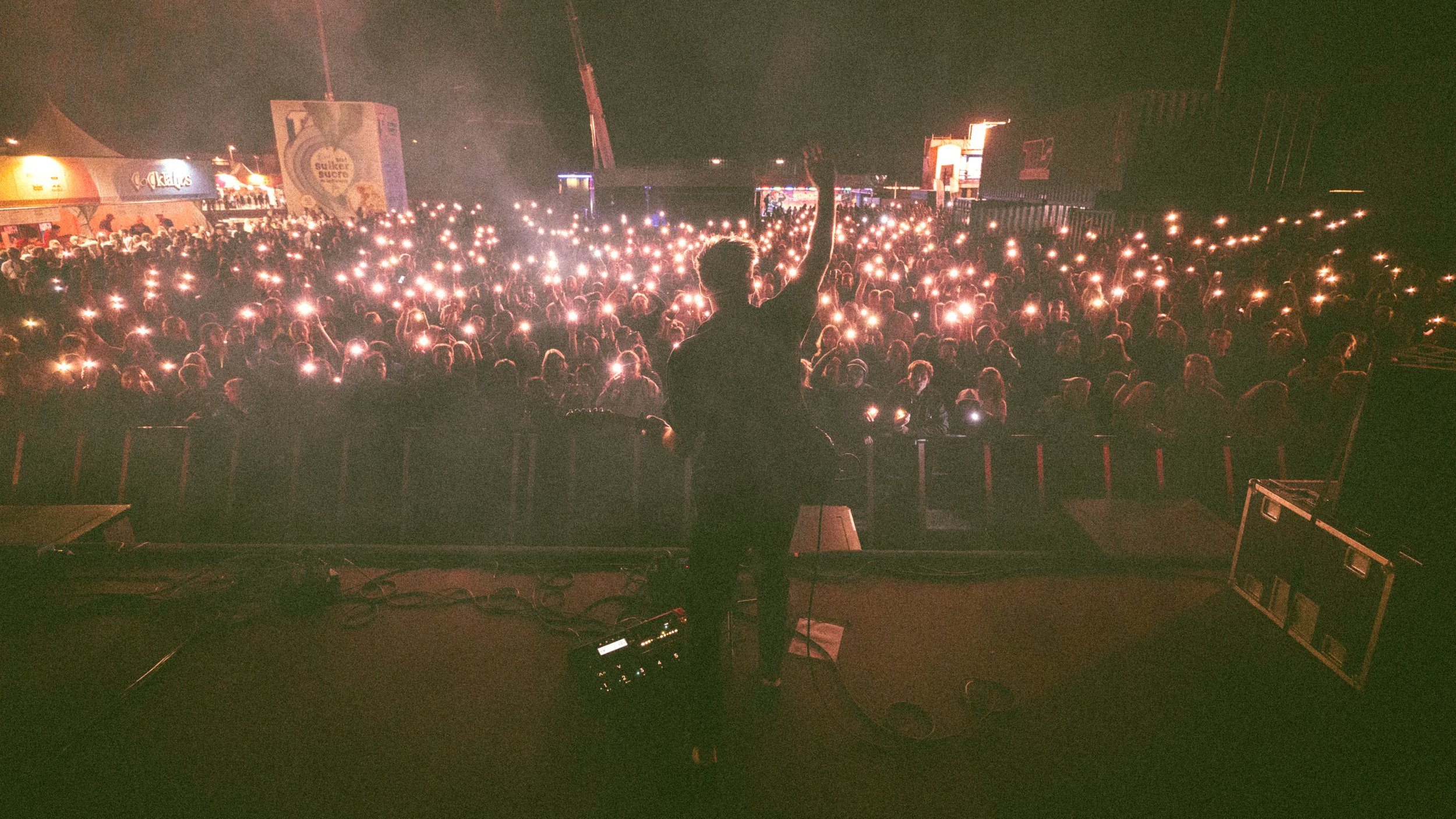 A DJ performing at night in front of a large crowd at a festival or concert, with many attendees holding up lights or phones creating a sparkling effect.