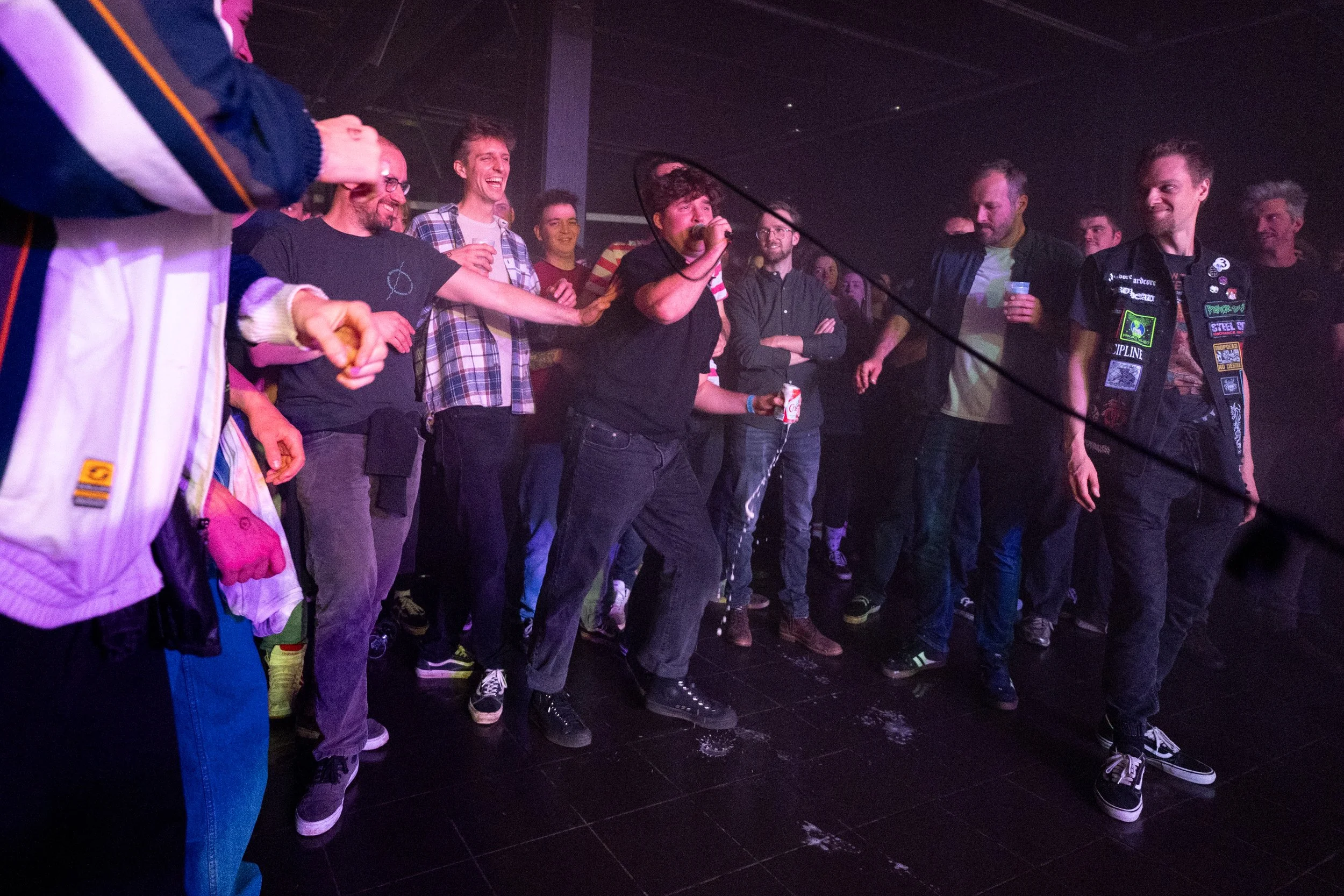 A group of people at a concert or party, with one person singing into a microphone while others around them are smiling, laughing, and holding drinks.